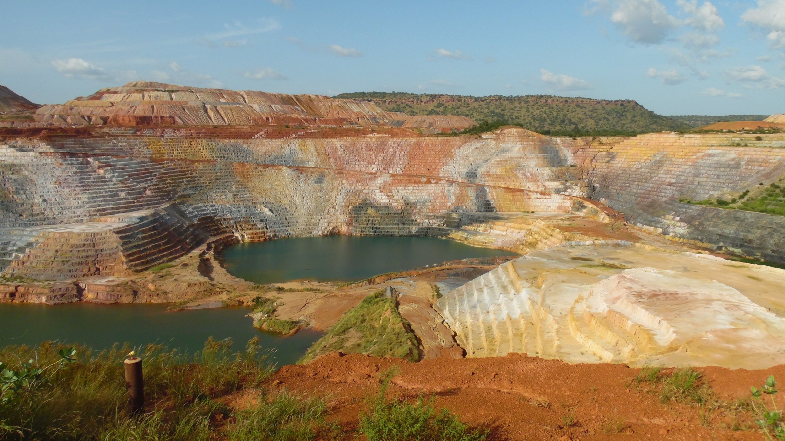 L'image montre une carrière de minerai, un paysage dramatique où les strates de terre créent des dégradés de couleurs allant du rouge au jaune et au blanc. Les parois de la carrière sont escarpées, formant des niveaux bien distincts. Au centre, un plan d'eau bleu émerge, contrastant avec les teintes terreuses et les paysages environnants. La végétation se fait rare sur les bords, mais quelques petites herbes vertes remontent le long des pentes. Le ciel est clair avec quelques nuages épars, ajoutant une lumière douce à la scène. Cette vue évoque à la fois l'exploitation humaine et la beauté naturelle brute.