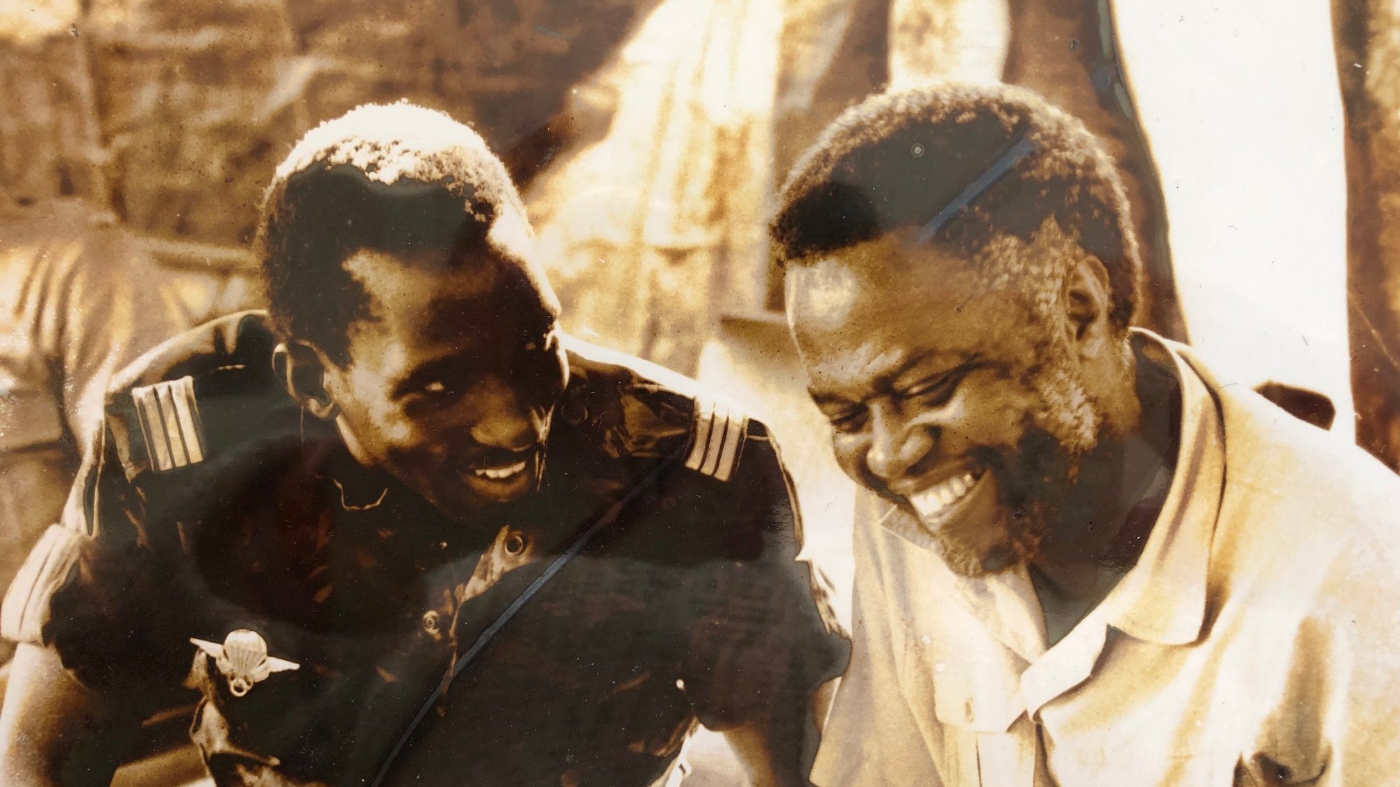 L'image montre deux hommes assis côte à côte, visiblement en train de discuter et de partager un moment de complicité. L'un des hommes porte un uniforme militaire, tandis que l'autre est vêtu d'une chemise claire. Ils affichent des sourires sincères, ce qui dénote une ambiance chaleureuse et amicale. L'arrière-plan est flou, mais on peut deviner une scène informelle, peut-être un rassemblement ou une réunion. La photo donne une impression de camaraderie et de joie partagée entre les deux individus.