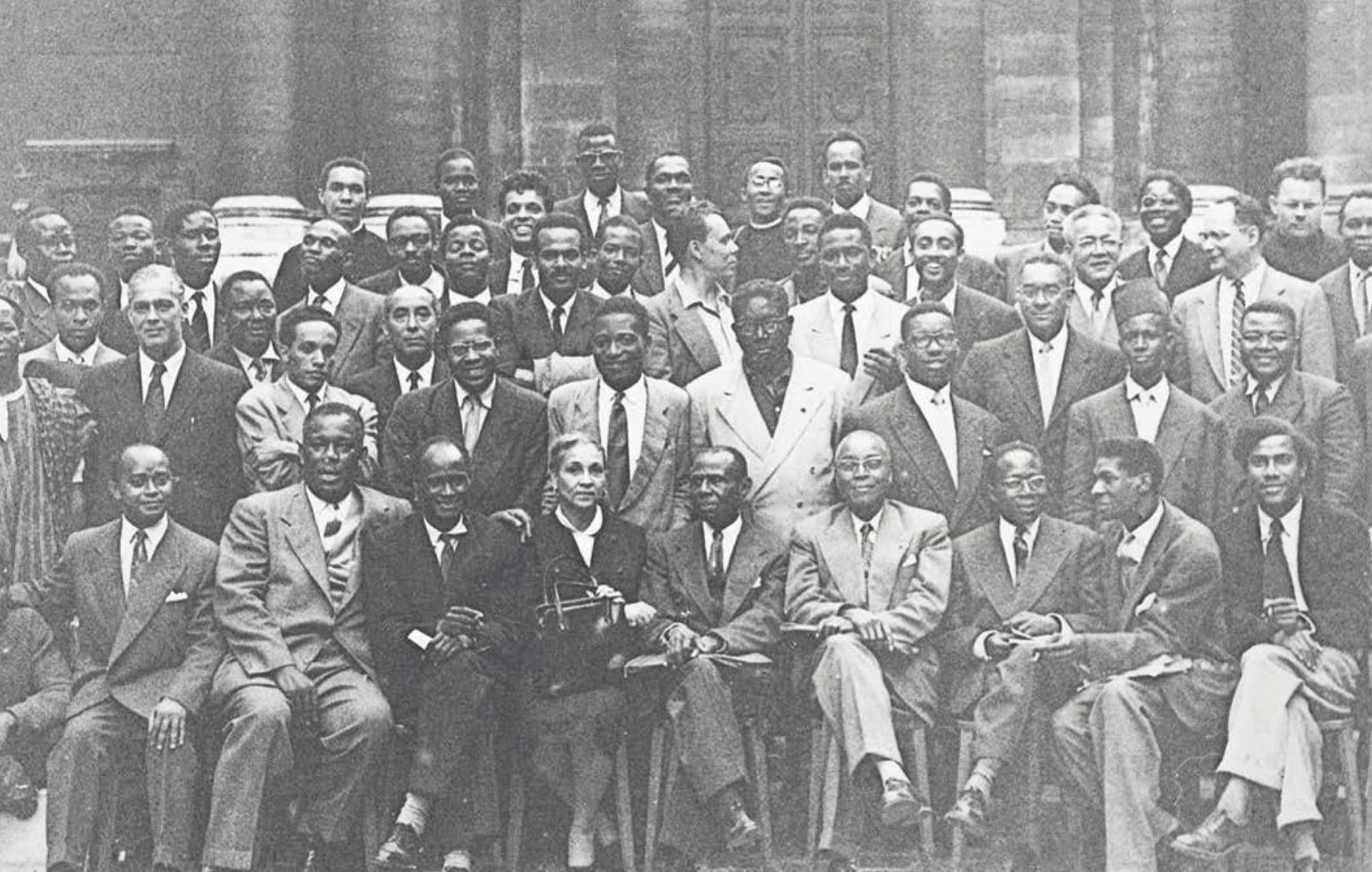 L'image présente un groupe de plusieurs hommes, probablement des leaders et des intellectuels, rassemblés pour une photographie de groupe. Ils sont vêtus de costumes formels, avec une variété de poses : certains sont assis, d'autres debout. L'ambiance semble chaleureuse et conviviale. Au centre, une femme se distingue, assise avec un sourire. En arrière-plan, on aperçoit un bâtiment historique, suggérant peut-être un événement ou une rencontre importante. L'image est en noir et blanc, ce qui lui donne un aspect intemporel.