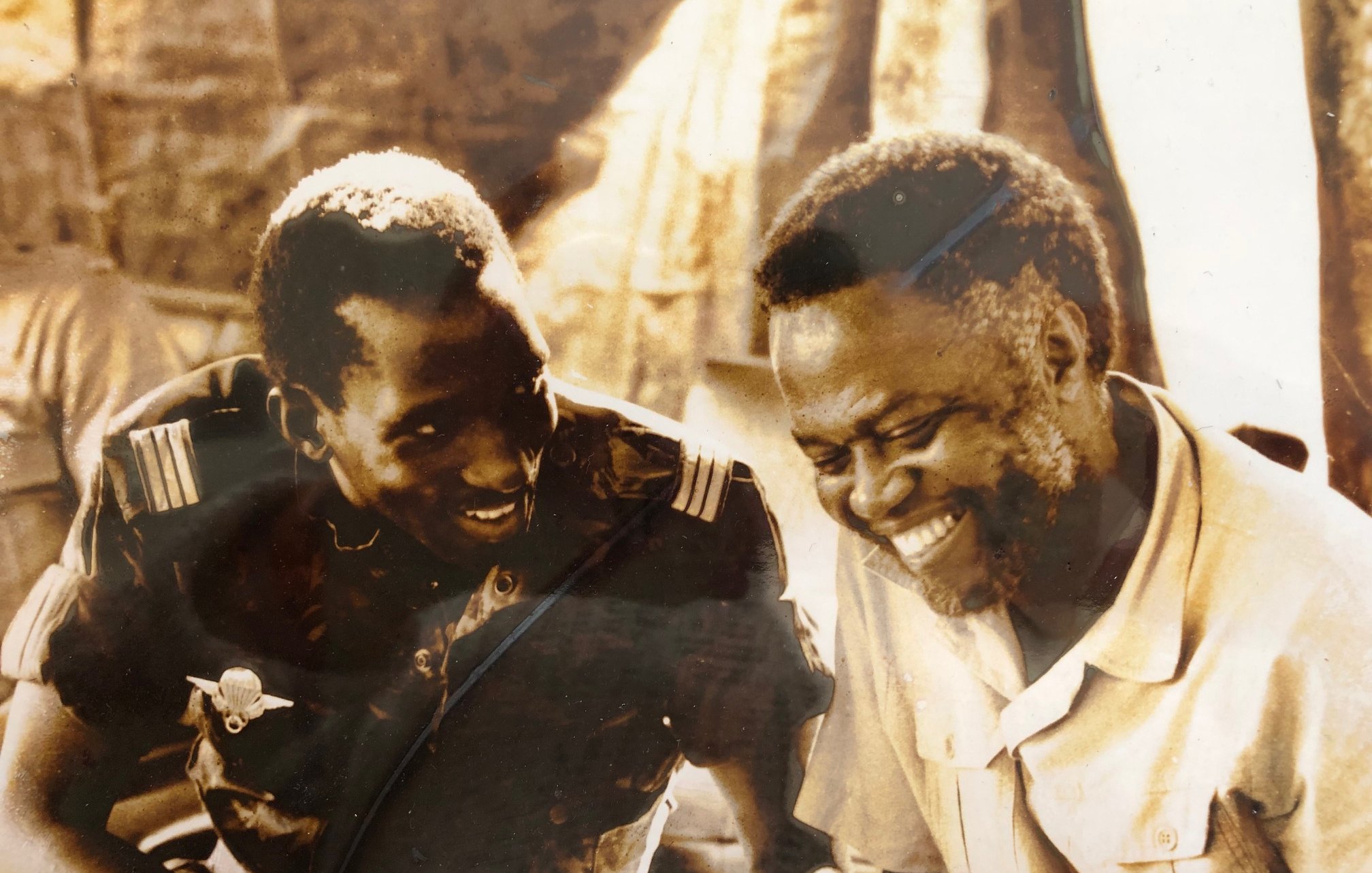 L'image montre deux hommes assis côte à côte, visiblement en train de discuter et de partager un moment de complicité. L'un des hommes porte un uniforme militaire, tandis que l'autre est vêtu d'une chemise claire. Ils affichent des sourires sincères, ce qui dénote une ambiance chaleureuse et amicale. L'arrière-plan est flou, mais on peut deviner une scène informelle, peut-être un rassemblement ou une réunion. La photo donne une impression de camaraderie et de joie partagée entre les deux individus.