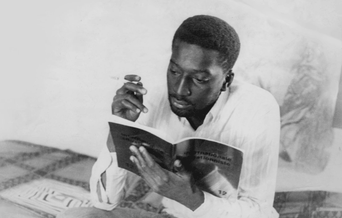 L'image montre un homme assis, plongé dans la lecture d'un livre. Il a la peau foncée et porte une chemise blanche. Dans sa main droite, il tient une cigarette. L'ambiance de la scène est calme, suggérant un moment de réflexion ou d'étude. L'arrière-plan évoque un espace intime, avec des motifs sur le sol qui peuvent donner une impression de confort. L'homme semble absorbé par sa lecture, dégageant une concentration et une sérénité.