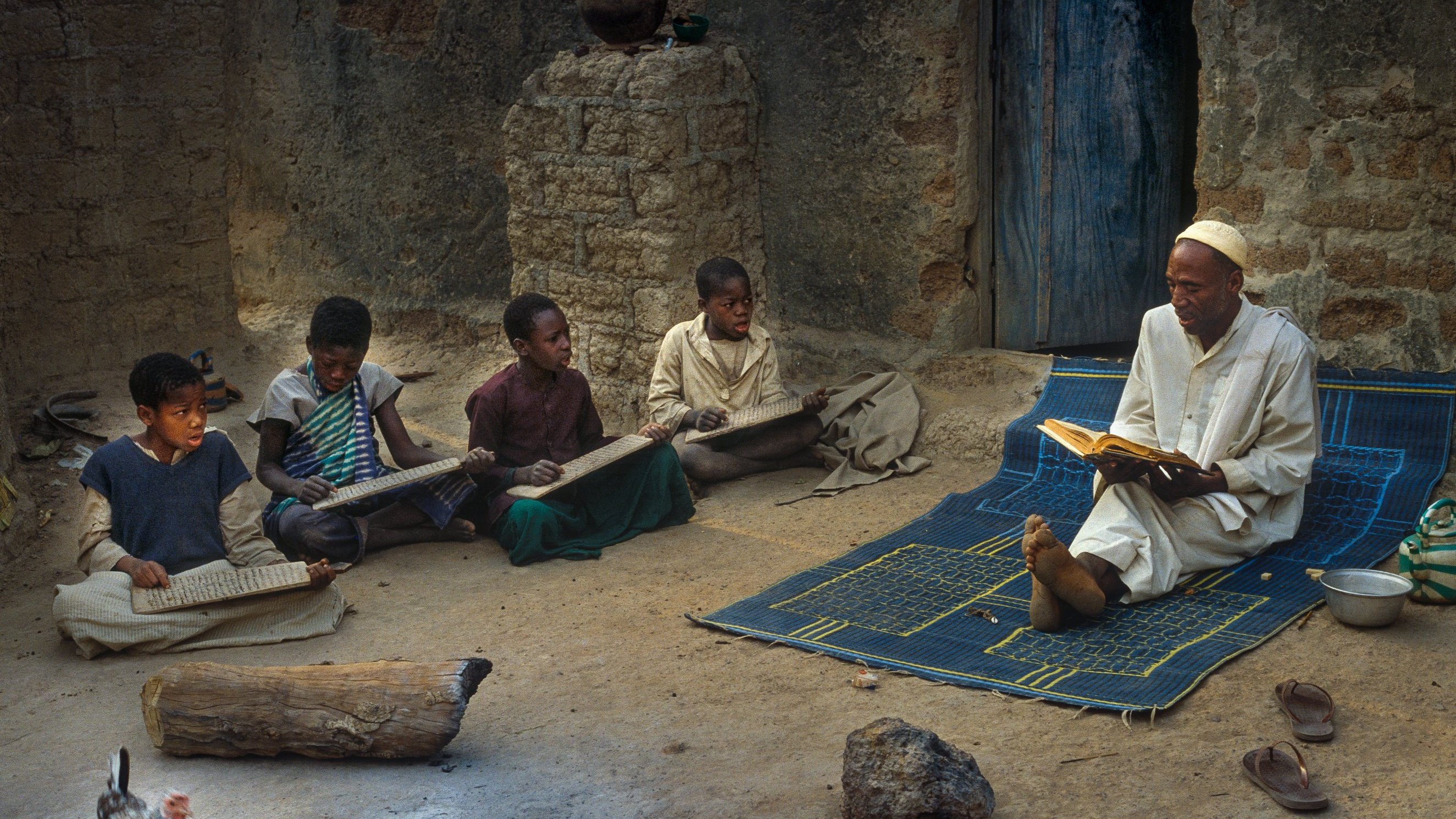 L'image montre un enseignant assis sur un tapis à l'extérieur d'une maison en terre. Il lit un livre avec attention, tandis que plusieurs enfants sont rassemblés autour de lui, concentrés sur des planches ou des livres qu'ils tiennent. L'ambiance est calme et studieuse. On peut aussi voir un poulet se déplacer au sol, ajoutant une touche de vie rurale à la scène. Les murs de la maison sont simples, et la lumière naturelle éclaire l'espace, créant une atmosphère chaleureuse et conviviale.