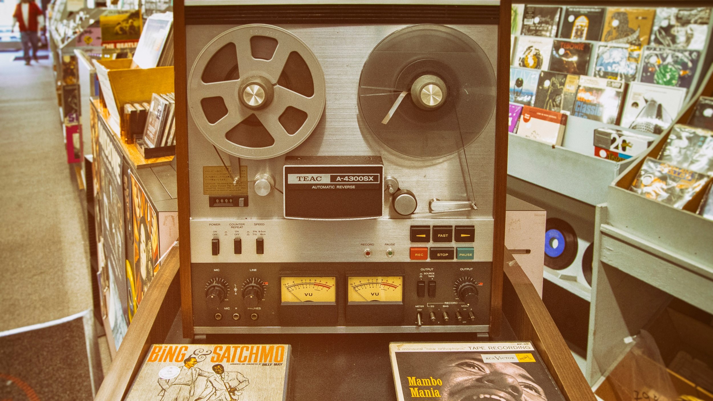 L'image montre un équipement audio vintage, un magnétophone à bande TEAC A-2300X, qui est posé sur un meuble en bois. Ce magnétophone a un design classique, avec un boîtier en métal argenté et des bobines de bande visibles sur le dessus. Deux compteurs analogiques sont situés en bas, indiquant les niveaux audio. À côté, on peut voir deux albums vinyles : l'un est intitulé "Bing & Satchmo", qui met en avant une collaboration entre le célèbre chanteur Bing Crosby et Louis Armstrong. L'autre album, "Mambo Mania" de Pérez Prado, suggère une atmosphère festive et rythmée. À l'arrière-plan, on aperçoit des étagères avec d'autres disques vinyles, créant une ambiance nostalgique et musicale.
