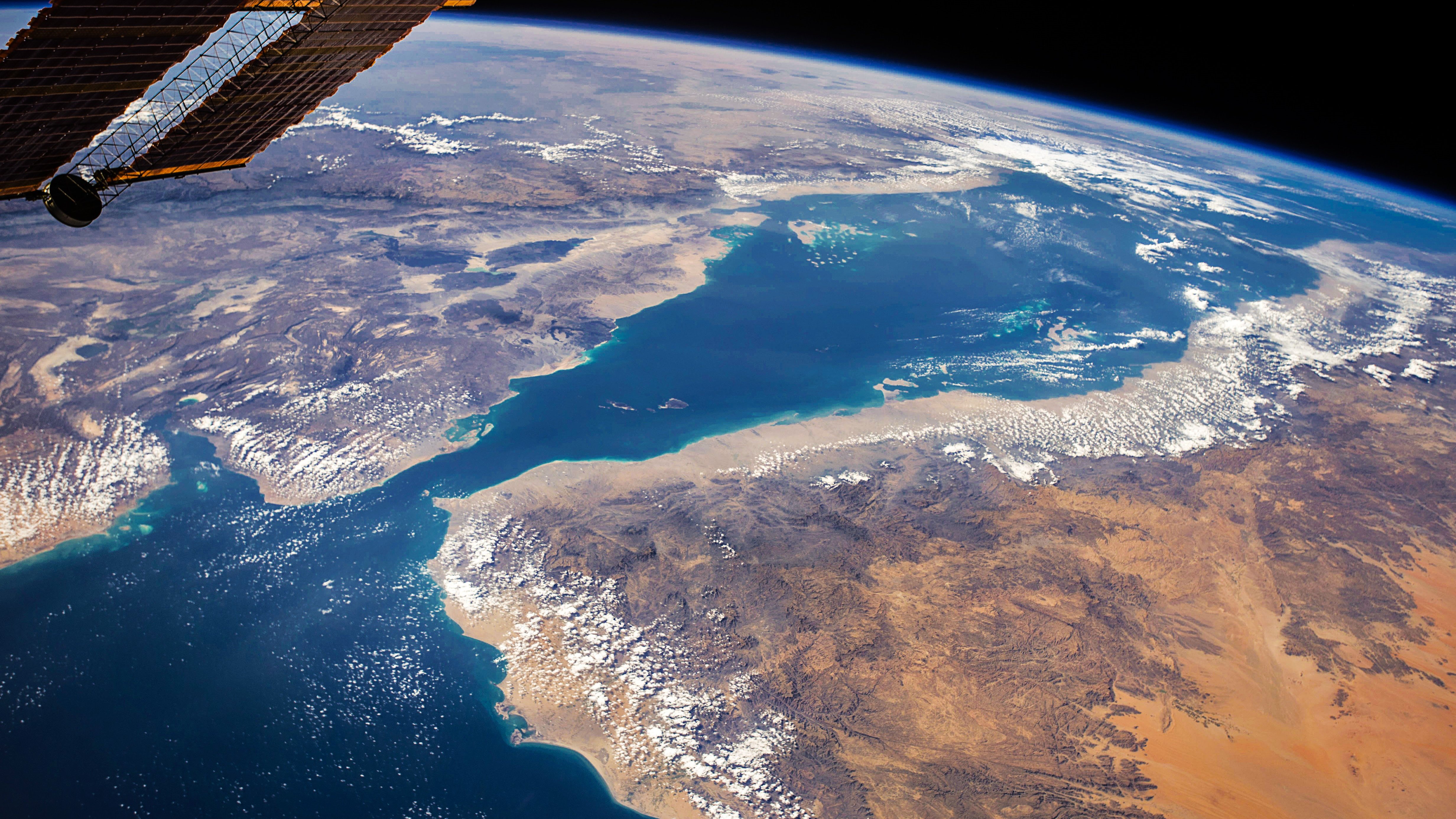 Cette image, prise depuis l'espace, montre une vue spectaculaire de la Terre. On y voit des paysages variés, avec une mer d'un bleu profond bordée de terres aux couleurs chaudes, tantôt sableuses, tantôt montagneuses. Il y a des nuages éparpillés qui ajoutent du relief à la scène. Les contours des côtes et des îles se dessinent nettement. En haut de l'image, on peut apercevoir la noirceur de l'espace, ce qui crée un beau contraste avec les teintes vives de la planète. Cette vue donne une impression de sérénité et d'immensité, rappelant la beauté fragile de notre environnement.