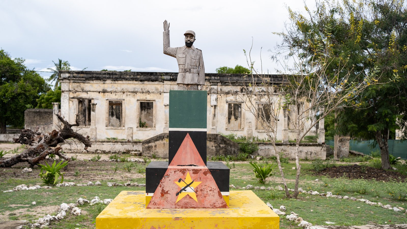 L'image montre un monument composé d'une statue d'un homme saluant, érigée sur une base colorée avec des formes géométriques. La statue est en métal et représente un personnage au regard déterminé. La base est peinte en plusieurs couleurs vives : rouge, jaune, vert et noir, formant des triangles. En arrière-plan, on aperçoit des ruines de bâtiments, avec des murs partiellement effondrés et une végétation verdoyante qui entoure l'ensemble. L'atmosphère est à la fois historique et contemplative, suggérant un lieu chargé de mémoire.