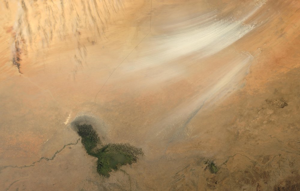 L'image montre une vaste étendue de paysage désertique, dominée par des teintes de beige et de marron clair. On aperçoit des stries blanches qui semblent indiquer des vents ou des mouvements de sable dans l'air. En bas de l'image, un petit plan d'eau apparaît, entouré de terres arides. Ce contraste entre la surface sèche du désert et l'eau brillante crée une scène à la fois aride et intrigante, témoignant des forces naturelles en jeu dans cet environnement désertique. Les textures et les couleurs évoquent une ambiance silencieuse et vaste, typique des paysages désertiques.
