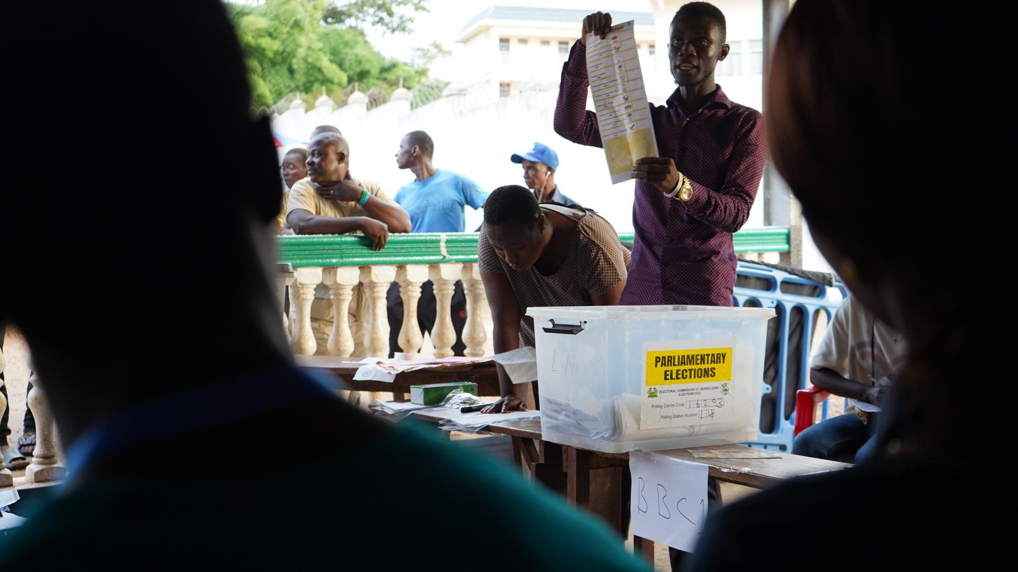 L'image montre une scène d'élections parlementaires. Au centre, un homme se tient debout, levant une feuille de papier, probablement un bulletin de vote, pour montrer aux personnes présentes. Il s'habille d'une chemise de couleur sombre et semble expliquer quelque chose de manière engageante. En arrière-plan, plusieurs personnes sont rassemblées, certaines observant attentivement tandis que d'autres attendent, adossées à une balustrade. On devine une atmosphère de concentration et d'anticipation, typique d'un moment électoral. Sur une table, une boîte transparente étiquetée "ELECTIONS PARLEMENTAIRES" est visible, contenant probablement des bulletins de vote.