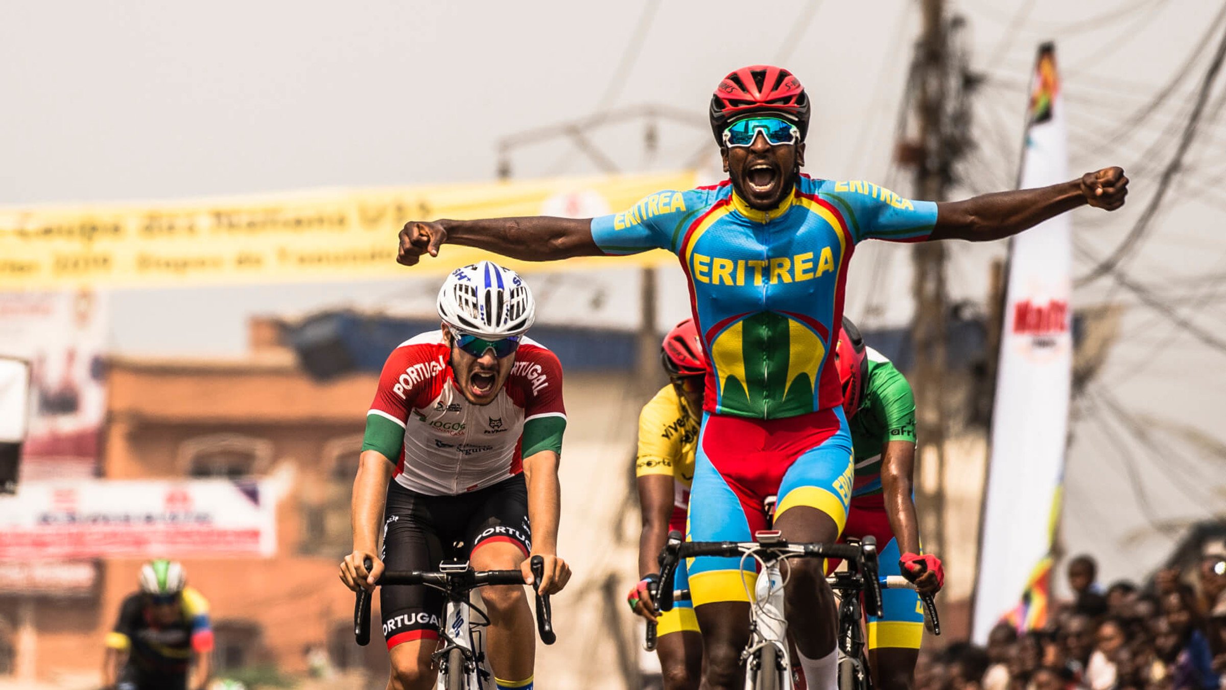 L'image montre un moment dynamique d'une course de cyclisme. Au premier plan, un coureur, vêtu d'un maillot multicolore représentant l'Érythrée, lève les bras en signe de victoire, affichant une expression de joie et d'excitation. En arrière-plan, un autre cycliste, portant un maillot aux couleurs du Portugal, semble montrer des signes d'effort intensif, presque à la limite de l'épuisement. On aperçoit également d'autres participants et une foule enjouée de spectateurs, créant une ambiance festive autour de cet événement sportif. Le décor est vibrant, évoquant l'énergie et la compétitivité de la course.
