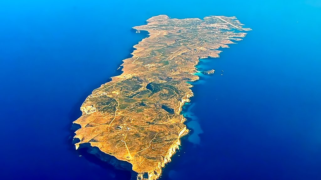 L'image représente une île vue du ciel, entourée par une vaste étendue d'eau bleue. L'île a une forme allongée et irrégulière, avec des côtes découpées et quelques zones rocheuses. Sa surface est en grande partie couverte de végétation, avec des teintes de jaune, de vert et de brun qui évoquent un terrain sec et accidenté. On peut également observer quelques petites baies et criques où l'eau semble se jeter sur le rivage. L'ensemble évoque une atmosphère de tranquillité et de beauté naturelle, typique d'une destination insulaire.
