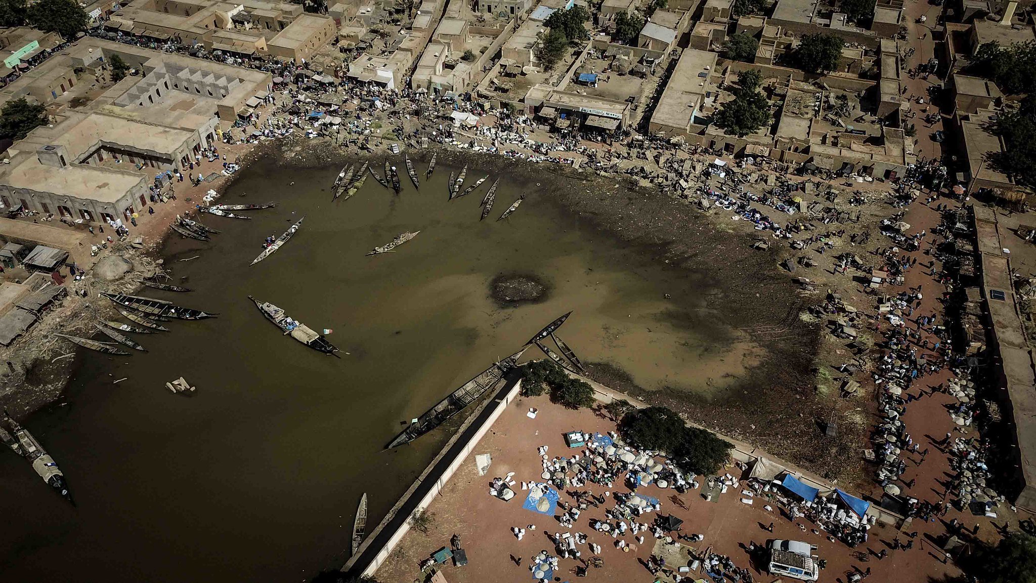 Imaginez un paysage vu du ciel. Au centre, un plan d'eau sombre qui semble être une lagune ou un port. Les bords de l'eau sont bordés de bateaux, certains amarrés, d'autres flottant paisiblement. Autour de cette lagune, il y a des habitations, des maisons en terre ou en tuiles, formant un petit village. Sur les rives, des gens sont rassemblés, certains assis par terre, d'autres se déplaçant avec des marchandises. Des tentes et des parasols sont disposés, suggérant un marché vivant où l'on troque et vend des produits. L'air est probablement rempli de sons, de cris et d'odeurs de nourriture, de poissons et de marchandises variées, donnant l'impression d'une scène animée et prospère. Les couleurs sont chaudes, évoquant un climat ensoleillé. Ce tableau dégage une atmosphère d'activité et de communauté autour de l'eau.