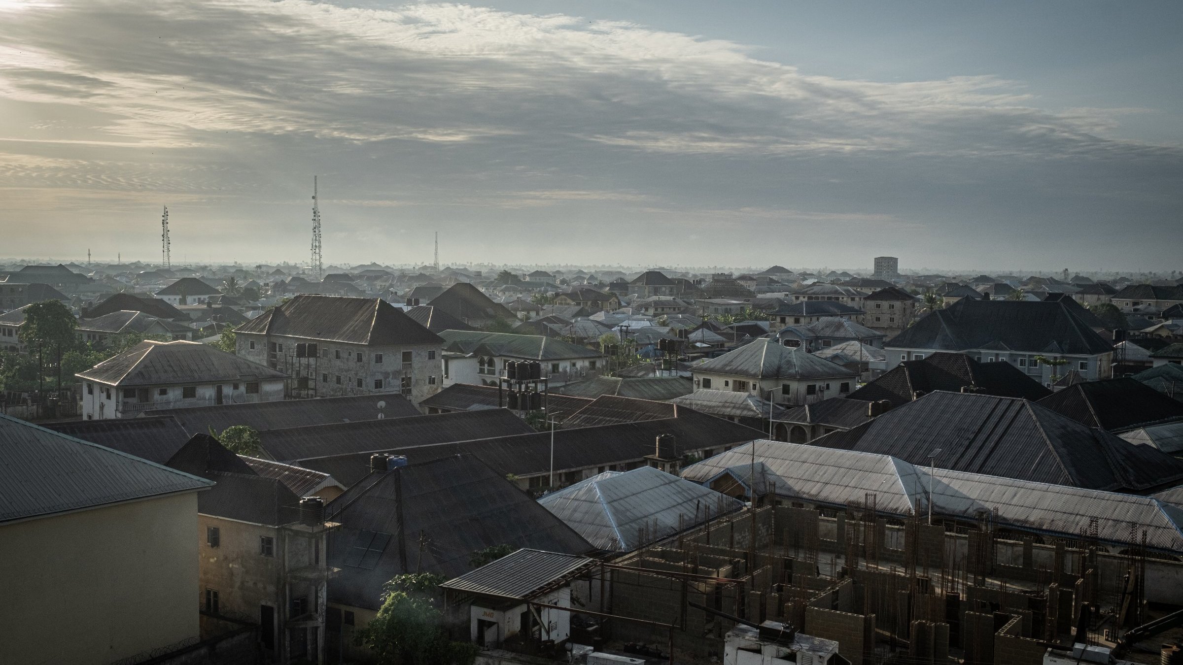 Dans cette image, on voit un paysage urbain d'une ville d'Afrique, probablement au lever du soleil. Les toits de différentes maisons se chevauchent, formant une mosaïque de formes et de textures. La majorité des toits sont en tôle, certains ayant des angles abrupts, tandis que d'autres sont plats. En arrière-plan, on aperçoit des constructions en cours, avec des structures en béton qui indiquent un développement urbain. Le ciel est parsemé de nuages, offrant une lumière douce et diffuse. On devine une atmosphère paisible au matin, avec des ombres légères qui accentuent les contours des bâtiments. Au loin, on distingue des antennes et des colonnes, signes de l'urbanisation moderne. L'ensemble dégage une impression de vie et d'activité, mais aussi de tranquillité à cette heure matinale.