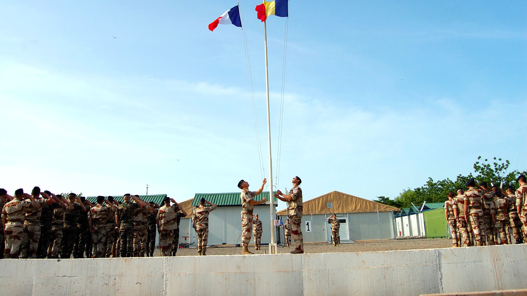 Cette image montre une cérémonie de lever de drapeaux dans un environnement militaire. Au premier plan, deux soldats se tiennent près d'un mât, où ils sont en train de hisser les drapeaux français et roumain. Ils sont parfaitement alignés et semblent respectueux. En arrière-plan, un groupe de soldats en uniforme assiste à la cérémonie, formant une ligne. L'environnement est clair et ensoleillé, avec des bâtiments militaires simples et une légère brise qui pourrait faire flotter les drapeaux. L'atmosphère est solennelle et empreinte de respect.
