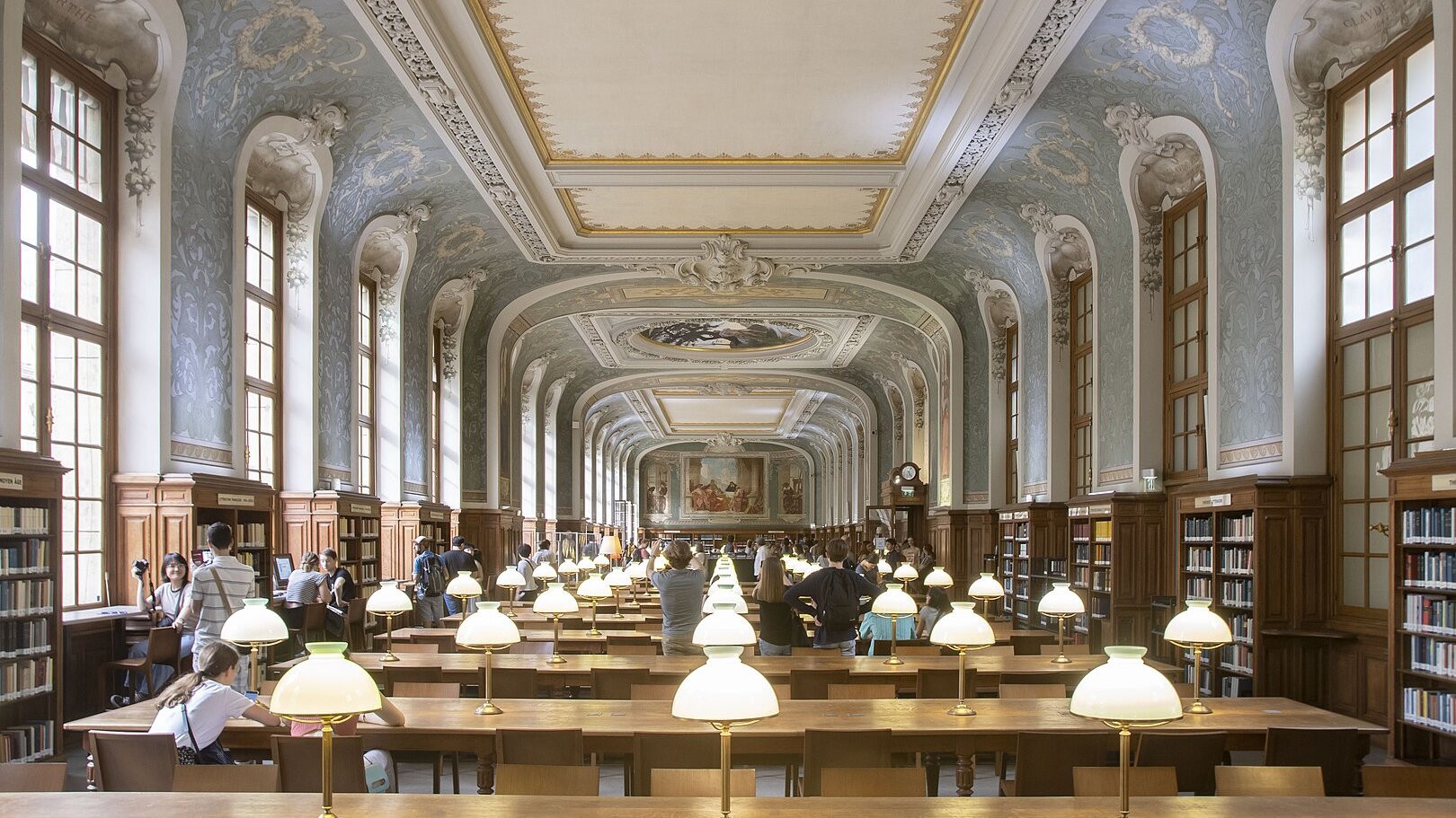 Imaginez une vaste bibliothèque, où l'air est chargé de silence et de connaissance. L'espace est long et majestueux, avec un plafond haut orné de décorations raffinées et de motifs artistiques. Les murs sont bordés de grandes fenêtres, permettant à la lumière naturelle d'inonder la pièce. Des lampes de table éclatent par leur lumière douce sur de longues tables en bois, disposées en rangées. Ces tables accueillent des visiteurs plongés dans leurs livres et leurs pensées. Au fond, un grand mur est décoré de fresques, ajoutant une note d'élégance à l'ensemble. Les étagères en bois, pleines de livres soigneusement rangés, s'élèvent majestueusement sur les côtés, évoquant un immense trésor de savoir à portée de main. Les gens se déplacent doucement, parlant à voix basse, ajoutant une ambiance de respect et de tranquillité. Cette bibliothèque semble être un sanctuaire pour les amateurs de lecture et de culture.