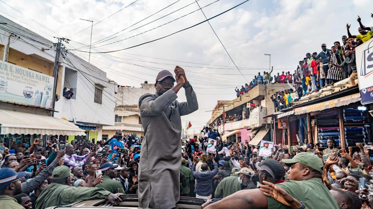 L'image montre une scène dynamique et animée dans une rue bondée. Au centre, un homme, vêtu d'une tenue grise, se tient debout sur un véhicule, levant les bras avec enthousiasme. Les gens autour de lui, une foule importante, applaudissent et expriment leur soutien. On peut entendre des cris de joie et des chants de la part des personnes qui entourent le véhicule. À l'arrière-plan, des bâtiments et des personnes sur les balcons ajoutent à l'atmosphère de ferveur et de célébration. Le ciel est partiellement nuageux, créant un éclairage intéressant sur cette scène vibrante.