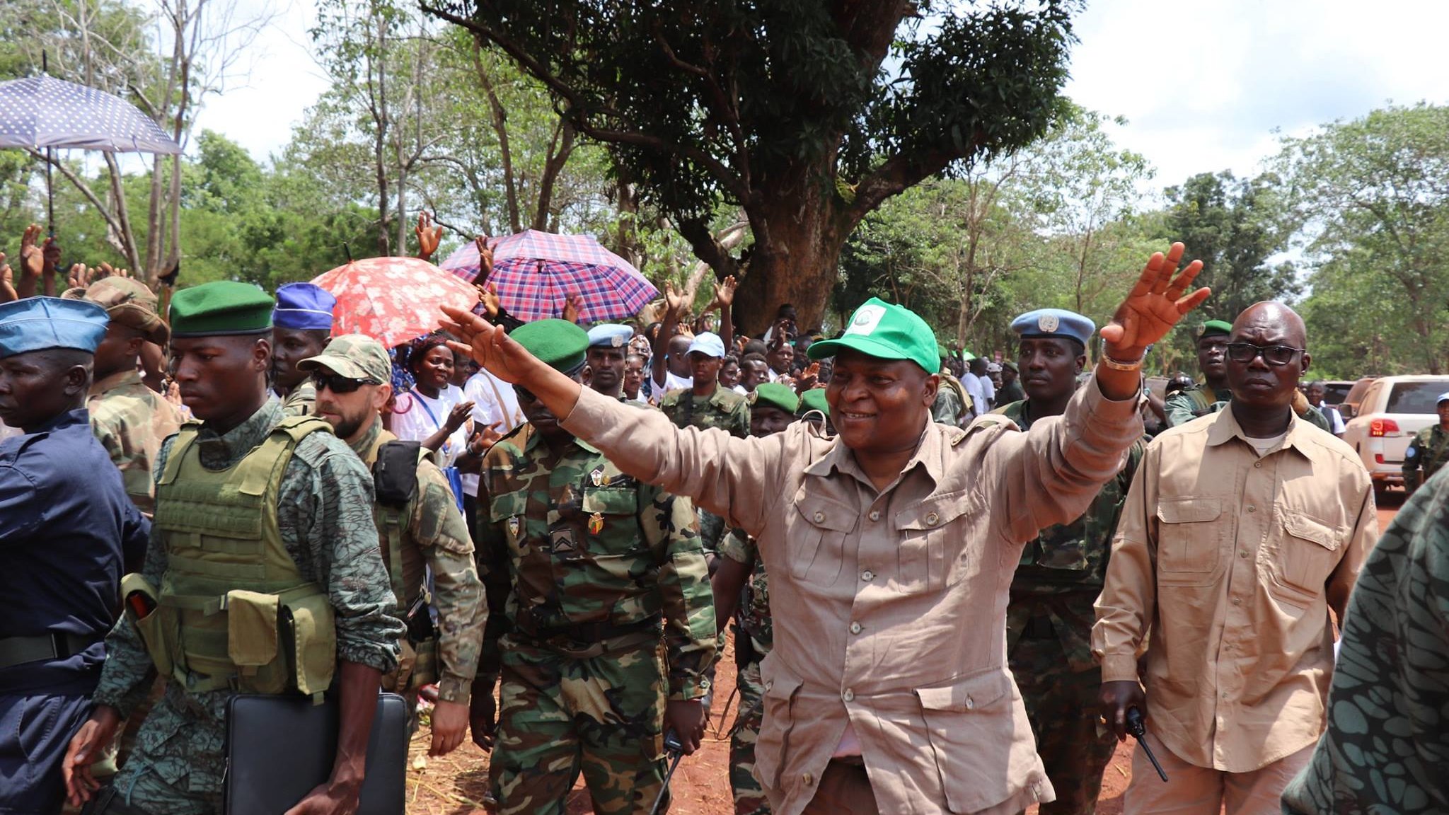 L'image montre une scène animée en plein air, avec un groupe de personnes réunies. Au centre, un homme souriant, vêtu d'une tenue beige et portant une casquette verte, semble saluer la foule avec les bras levés. Autour de lui, plusieurs militaires en uniforme, certains portant des gilets pare-balles, se tiennent en rang. La foule en arrière-plan est composée de personnes sous des parapluies, suggérant une atmosphère festive ou de célébration. Des arbres verdoyants et un chemin de terre sont visibles, créant un cadre naturel et vivant. L'ambiance semble énergique et encourageante.