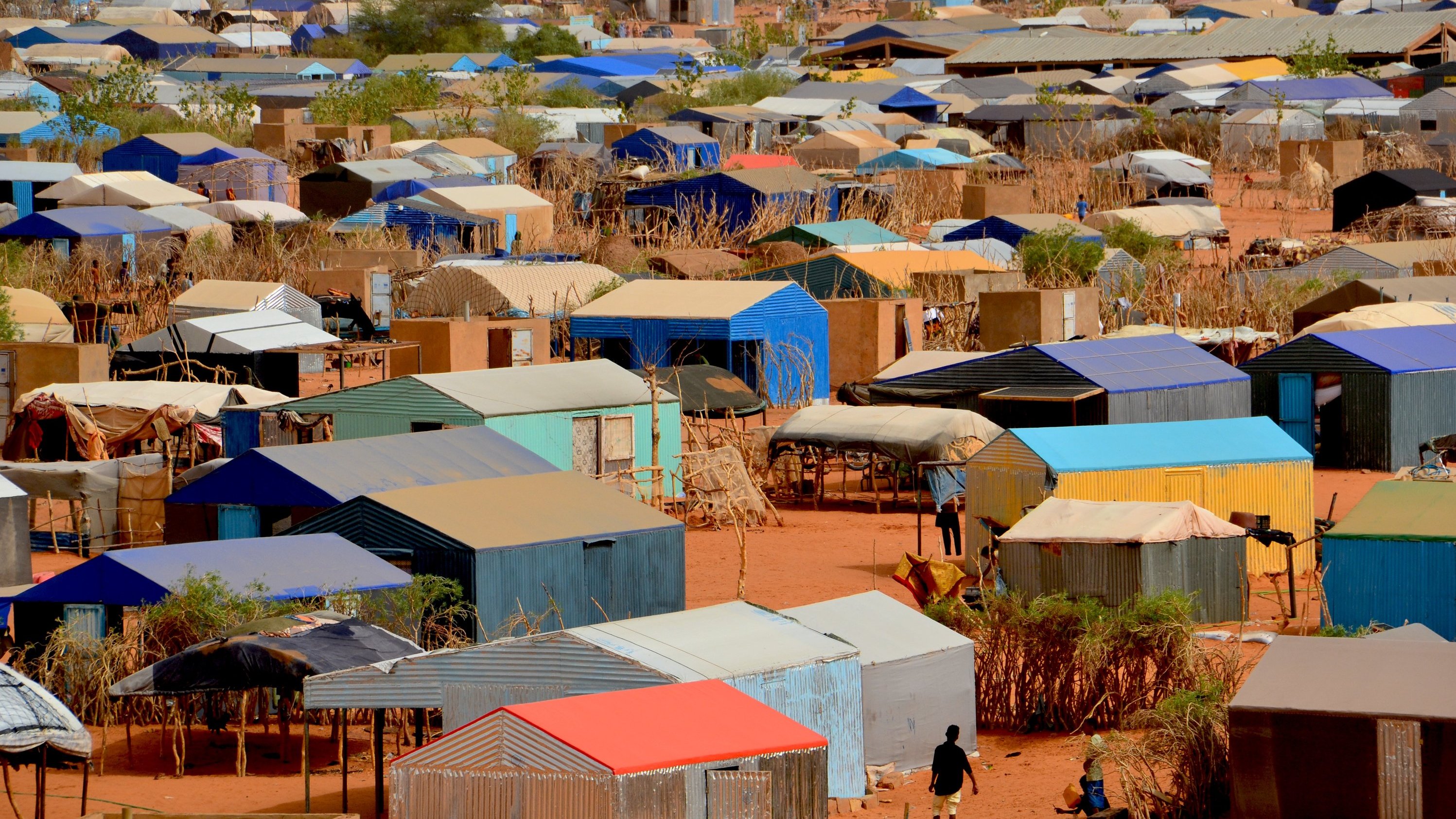 L'image montre un vaste camp de tentes et de logements temporaires disposés de manière dense sur un terrain désertique. Les structures sont recouvertes de toits de différentes couleurs, principalement des teintes de bleu, de rouge et de gris. Le sol est sablonneux, et quelques arbres ou buissons apparaissent ici et là, apportant un peu de verdure. En bas de l'image, on peut apercevoir des personnes se déplaçant entre les tentes, évoquant une activité quotidienne dans ce milieu. L'ensemble dégage une impression d'organisation dans la diversité des abris, tandis que le paysage aride souligne les conditions de vie dans ce camp.