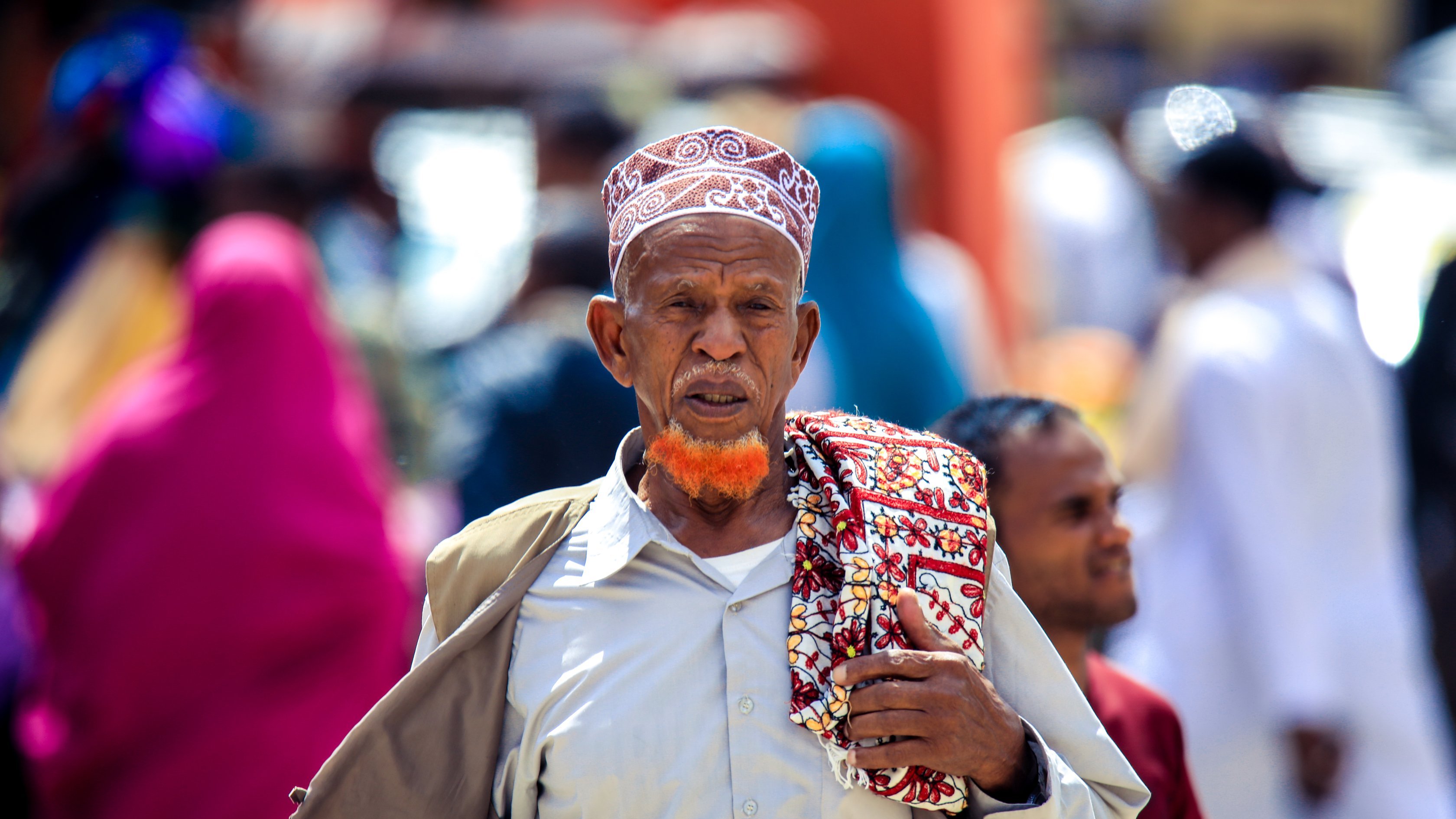 L'image montre un homme d'un certain âge marchant dans une rue animée. Il porte une tenue traditionnelle, avec une chemise claire et un grand châle coloré drapé sur son épaule. Sa tête est couverte d'un bonnet à motifs distinctifs, et il a une barbe teintée en orange, qui attire l'attention. En arrière-plan, on aperçoit des silhouettes floues de personnes vêtues de vêtements colorés, créant une ambiance vivante et dynamique. Les couleurs et la diversité des tenues témoignent d'un environnement culturel riche.