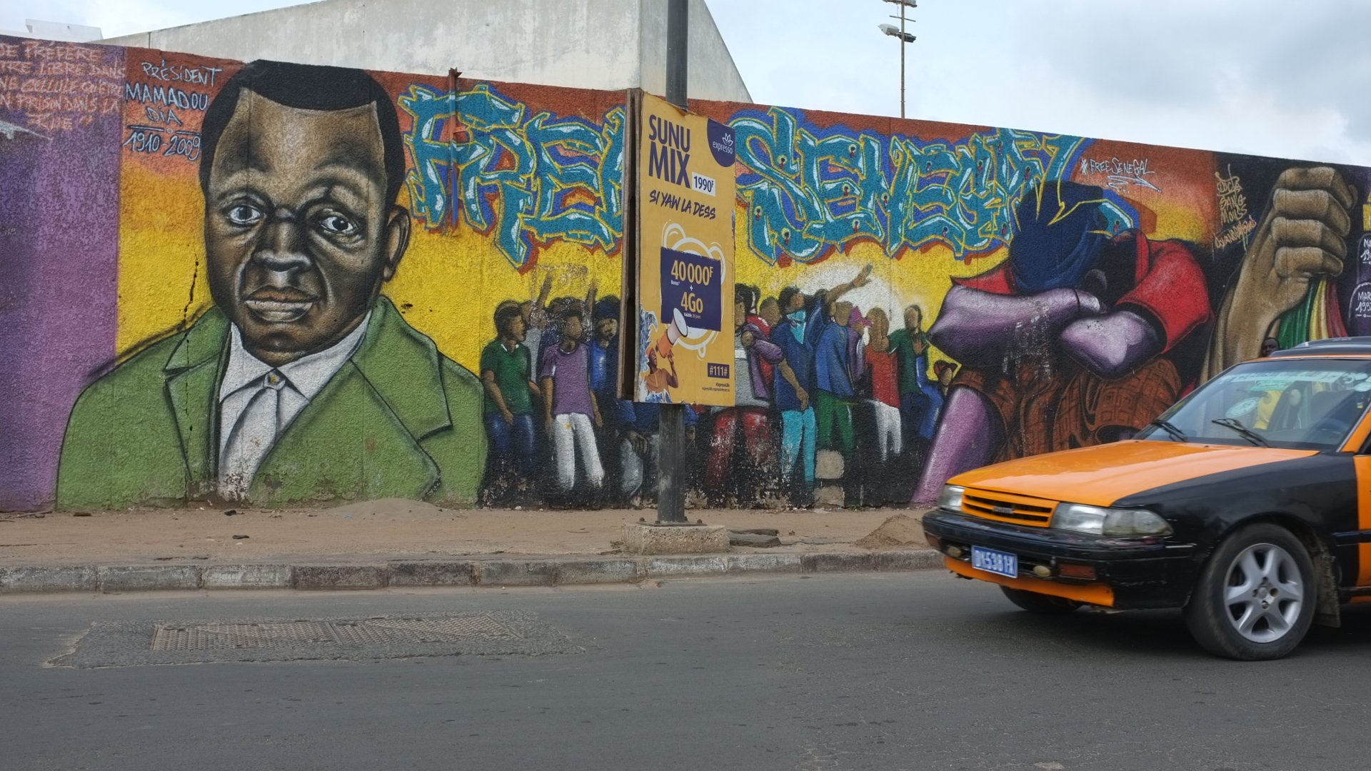 L'image montre un mur de graffitis vibrant et coloré. Au centre, il y a un portrait d'une personne au visage sérieux, avec des traits marqués, habillée d'un costume vert. Autour de ce portrait, différentes figures sont représentées, suggérant une scène animée de personnes rassemblées, peut-être en train de discuter ou de célébrer. À côté, on peut voir une affiche collée sur le mur avec des éléments graphiques. En bas de l'image, un taxi à la carrosserie orange et noir passe sur la route, ajoutant une touche de dynamisme à la scène urbaine. L'ensemble évoque une ambiance de vie communautaire et de culture urbaine.