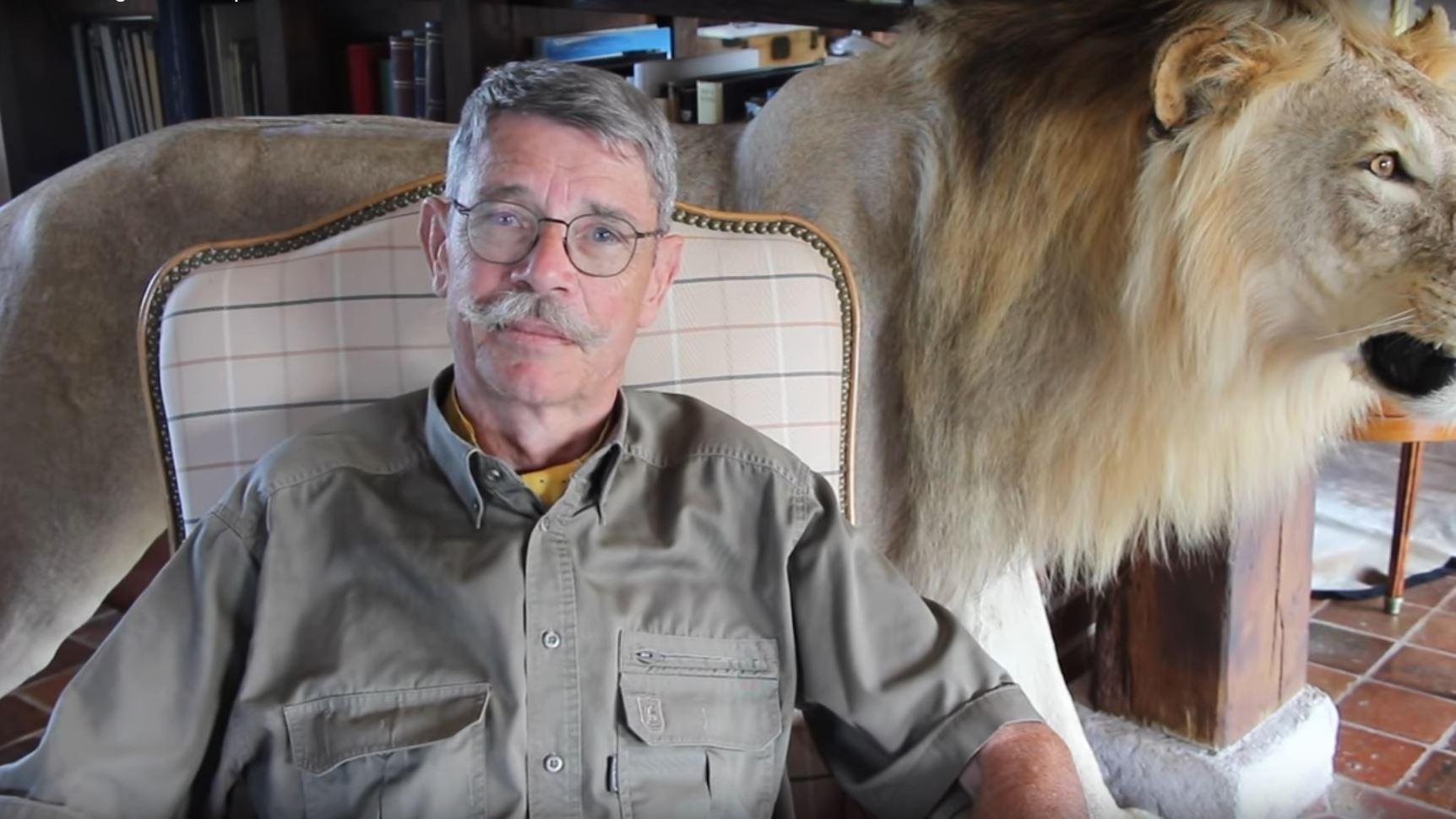 L'image montre un homme assis sur un fauteuil. Il a des lunettes, une moustache grise et porte une chemise kaki. À côté de lui, il y a un lion naturalisé, avec une crinière fournie et un regard majestueux. Le cadre donne une impression chaleureuse et rustique, avec une ambiance intérieure qui suggère une maison ou un chalet. Le contraste entre l'homme et le lion évoque une connexion avec la nature sauvage.