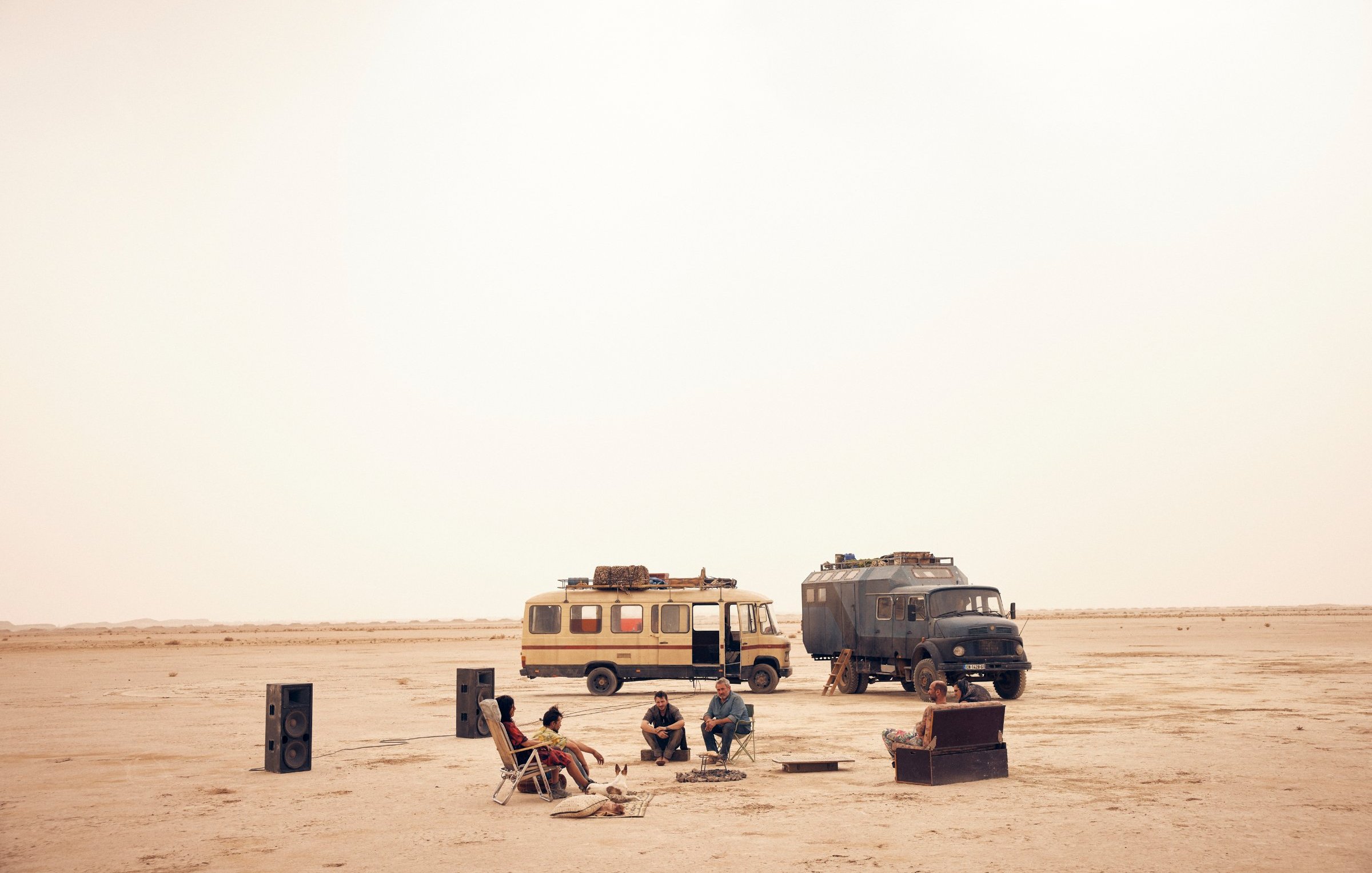 L'image montre un paysage désertique, vaste et aride, baigné par une lumière douce. Au centre, un groupe de personnes est assis autour d'un feu de camp. Ils semblent se détendre et passer un bon moment ensemble. À proximité, deux véhicules distincts attirent l'attention : l'un est un camping-car jaune, l'autre un véhicule utilitaire de couleur sombre. Des haut-parleurs ont été disposés autour du groupe, évoquant une ambiance festive. Le sol est sablonneux, et l'atmosphère semble calme, presque magique, sous un ciel d'une teinte uniforme.