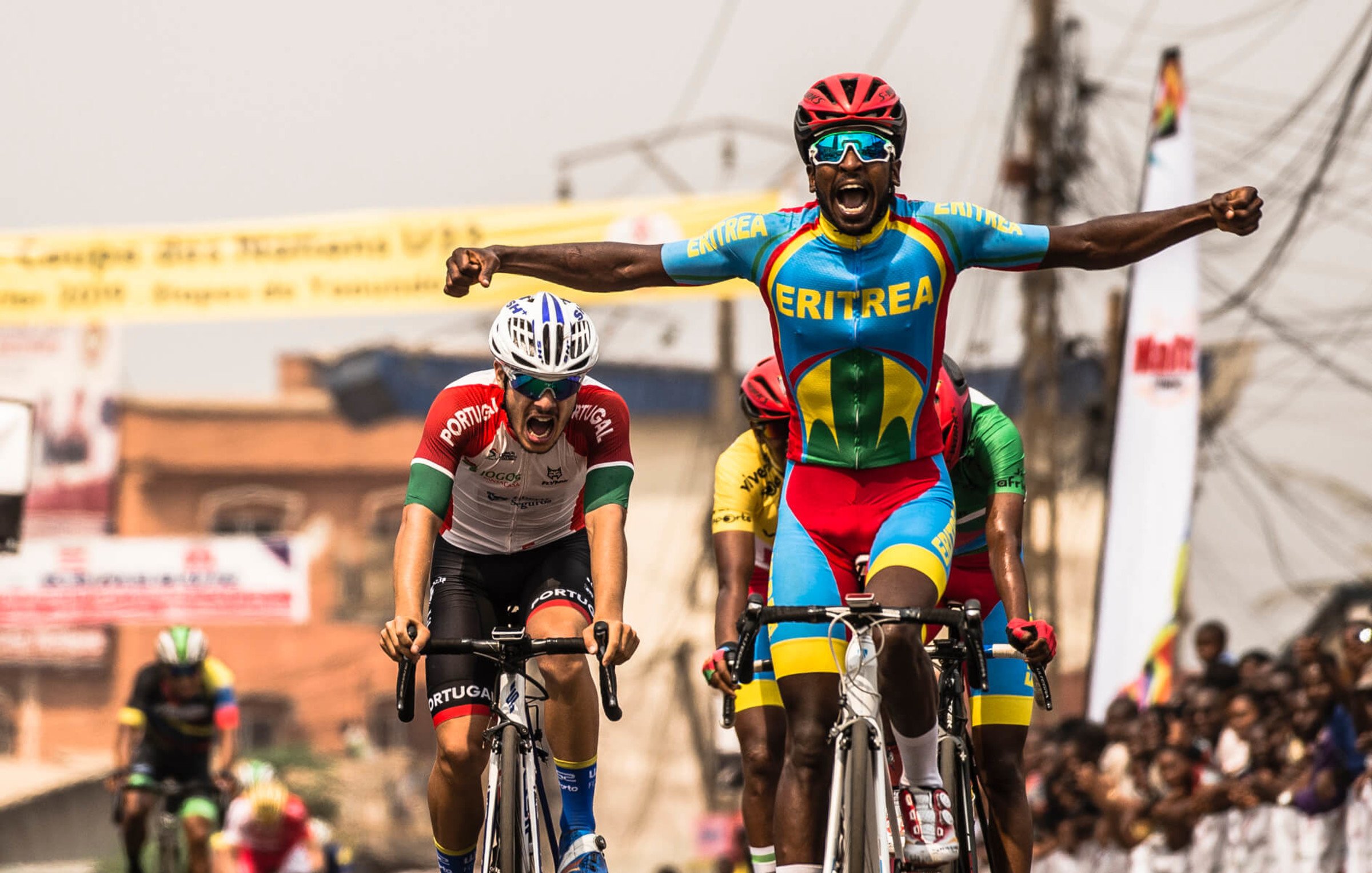 L'image montre un moment dynamique d'une course de cyclisme. Au premier plan, un coureur, vêtu d'un maillot multicolore représentant l'Érythrée, lève les bras en signe de victoire, affichant une expression de joie et d'excitation. En arrière-plan, un autre cycliste, portant un maillot aux couleurs du Portugal, semble montrer des signes d'effort intensif, presque à la limite de l'épuisement. On aperçoit également d'autres participants et une foule enjouée de spectateurs, créant une ambiance festive autour de cet événement sportif. Le décor est vibrant, évoquant l'énergie et la compétitivité de la course.