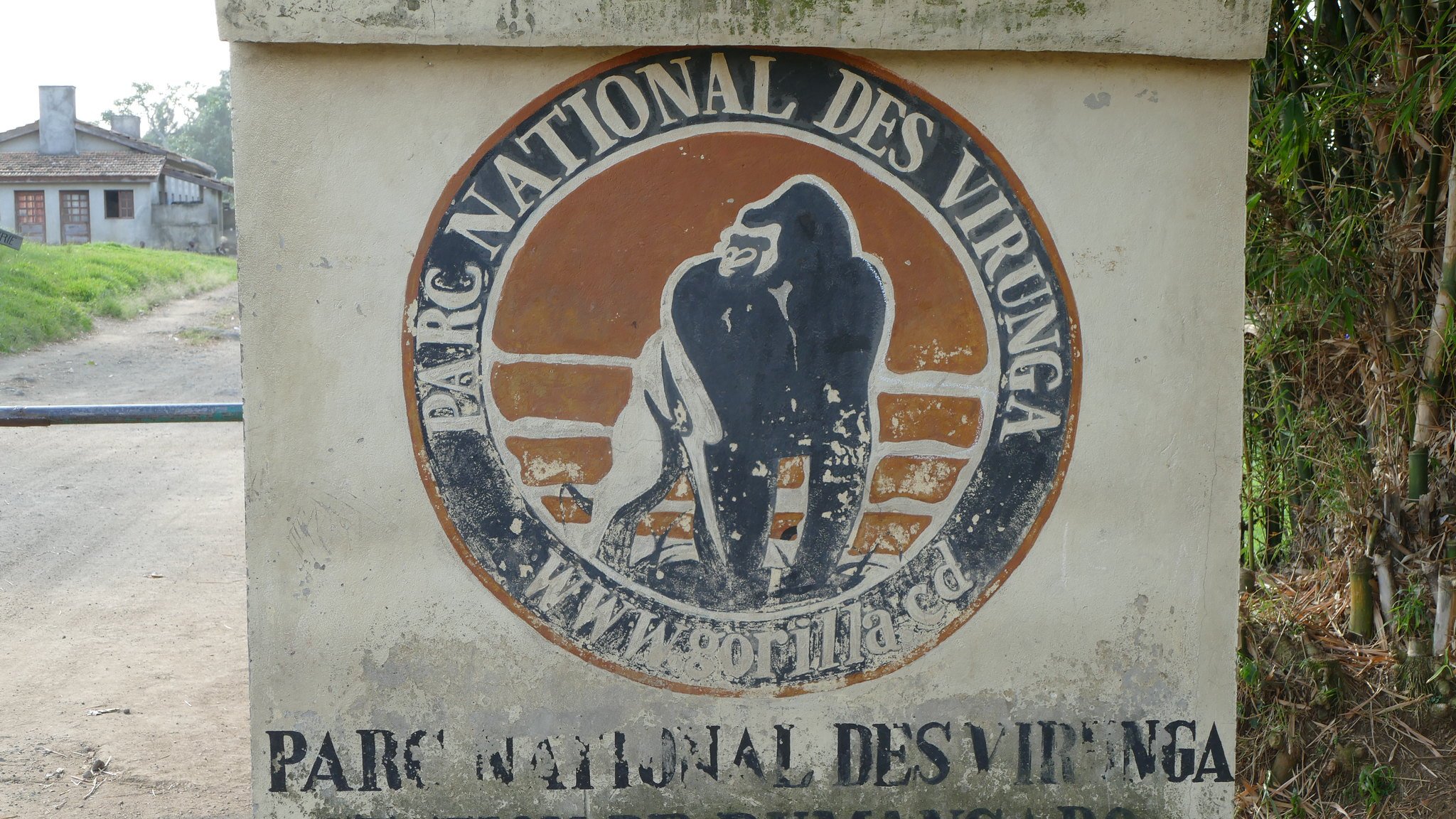 L'image montre un panneau en béton qui indique l'entrée du Parc National des Virunga. Le panneau est décoré d'un logo représentant un gorille, qui est l'un des emblèmes du parc. Les mots "PARC NATIONAL DES VIRUNGA" et "STATION DE RUMANGABO" sont inscrits en lettres claires sur la surface du panneau. En arrière-plan, on peut apercevoir des bâtiments et une route qui semble mener vers la nature environnante. L'ensemble de la scène évoque une atmosphère d'aventure et de préservation de la faune.