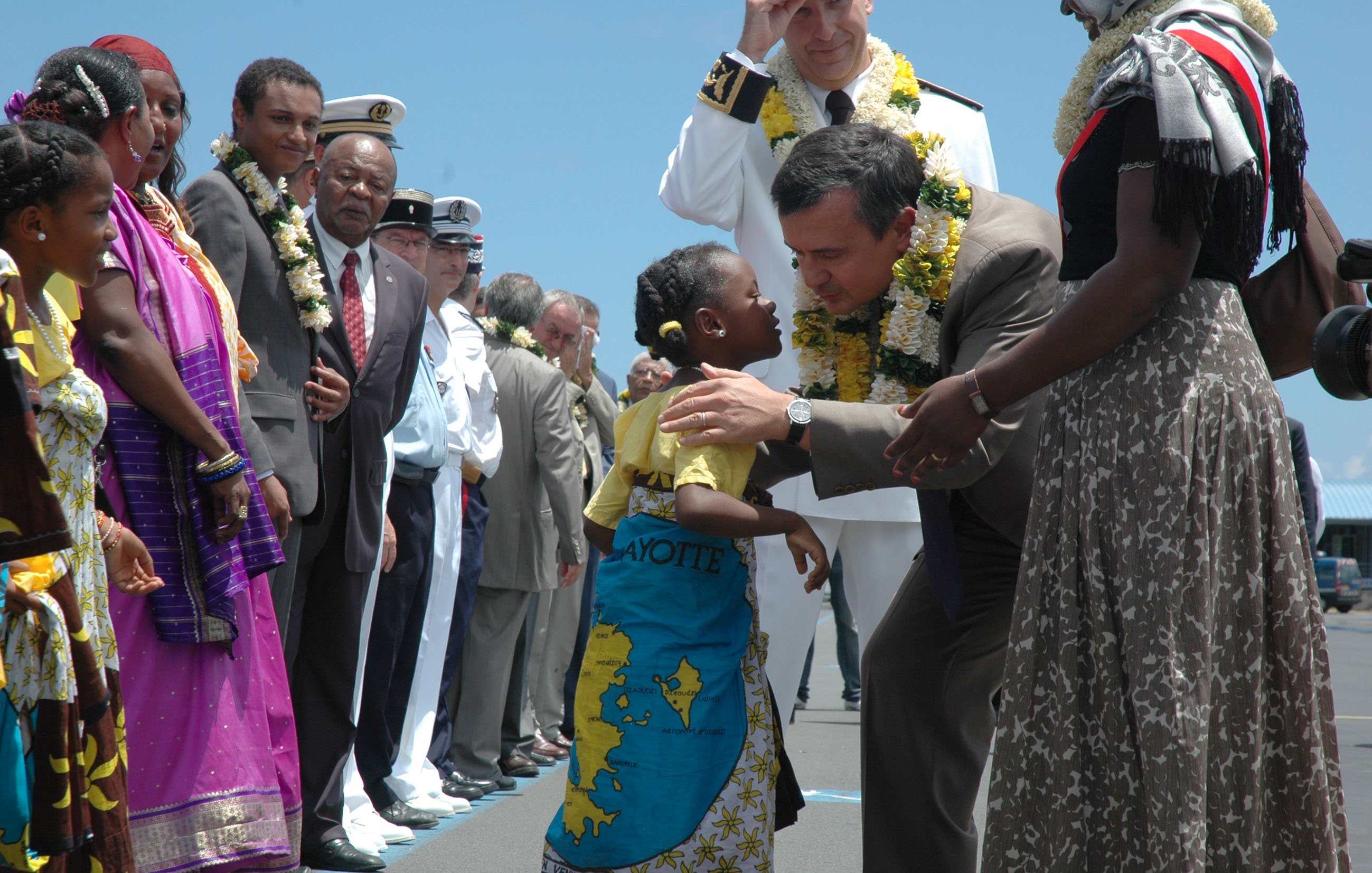 L'image représente une scène d'accueil vibrant sur un aéroport. Au premier plan, un homme en costard se penche pour embrasser une petite fille qui porte une robe colorée et un paréo. La fillette a un grand sourire et semble très joyeuse. Autour d'eux, plusieurs autres personnes, dont des adultes en vêtements traditionnels et militaires, les observent avec des expressions bienveillantes. Certains présents portent des guirlandes de fleurs autour du cou, ajoutant une touche festive à l'atmosphère. En arrière-plan, le ciel est clair, suggérant une journée ensoleillée. L'ensemble de la scène dégage une impression de chaleur et de convivialité.