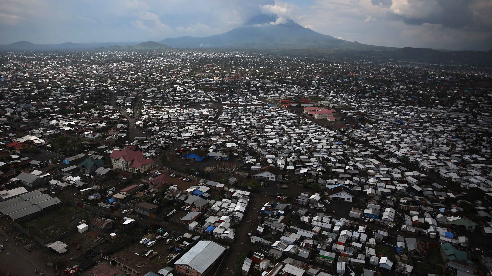 Imaginez une vaste étendue urbaine vue d'en haut, où des milliers de toits en tôle ondulée et en matériaux divers se mêlent pour créer un paysage dense. Les bâtiments semblent serrés les uns contre les autres, formant un réseau labyrinthique. Au loin, un majestueux volcan émerge à l'horizon, avec des sommets nuageux qui soulignent sa présence imposante. Le ciel présente des teintes variées de gris et de bleu, projetant une lumière diffuse sur la scène. En bas, des routes sont visibles, animées de véhicules et de personnes, ajoutant une sensation de vie à cet environnement. Cette image évoque à la fois la densité urbaine et la force tranquille de la nature en toile de fond.
