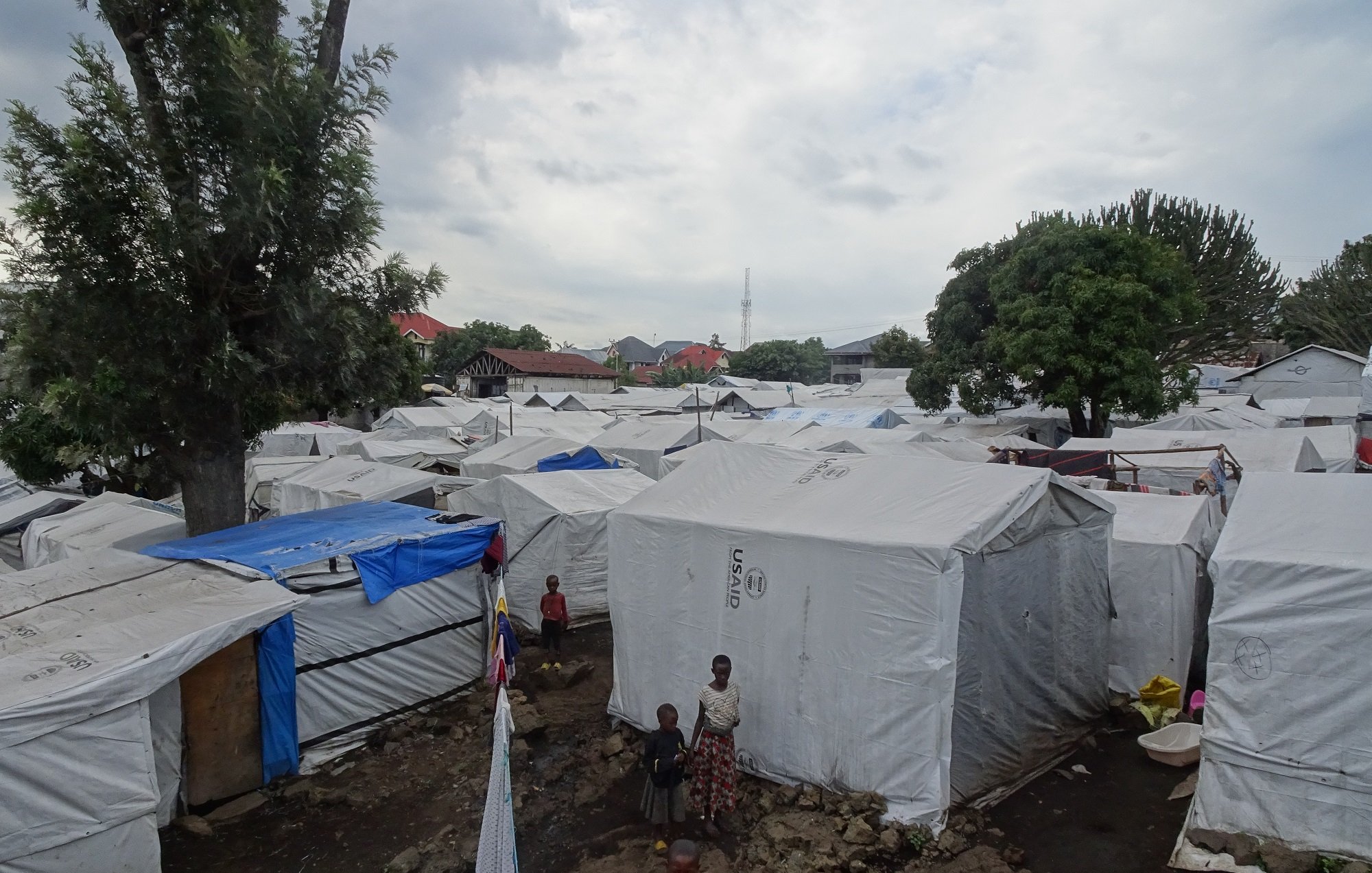 Cette image montre un paysage d'un camp de réfugiés, avec de nombreuses tentes blanches disposées de manière un peu désordonnée. Les tentes sont recouvertes d'une toile claire qui les protège des intempéries. On aperçoit quelques enfants jouant au milieu des tentes. Au loin, des arbres abondants ajoutent une touche de verdure, et on peut distinguer quelques structures plus solides. Le ciel est nuageux, suggérant qu'il pourrait pleuvoir, ce qui souligne la vulnérabilité du cadre. L'ambiance générale semble à la fois animée et calme, tandis que le sol est irrégulier, avec des pierres et des zones boueuses.