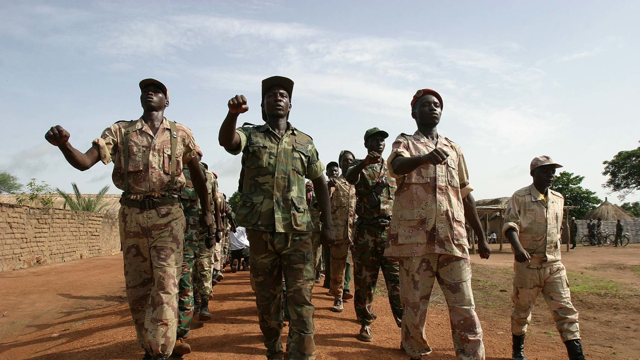 L'image montre un groupe de soldats marchant en formation sur un sol poussiéreux. Ils portent des uniformes militaires variés, allant du camouflé au beige, qui suggèrent une ambiance militaire. Certains soldats brandissent le poing en signe de détermination, tandis que d'autres avancent d'un pas ferme. En arrière-plan, on peut voir des tentes militaires ou des structures rudimentaires, suggérant un camp. Le ciel est légèrement nuageux, laissant passer quelques rayons de soleil. L'atmosphère générale évoque la discipline et la camaraderie au sein de ce groupe.