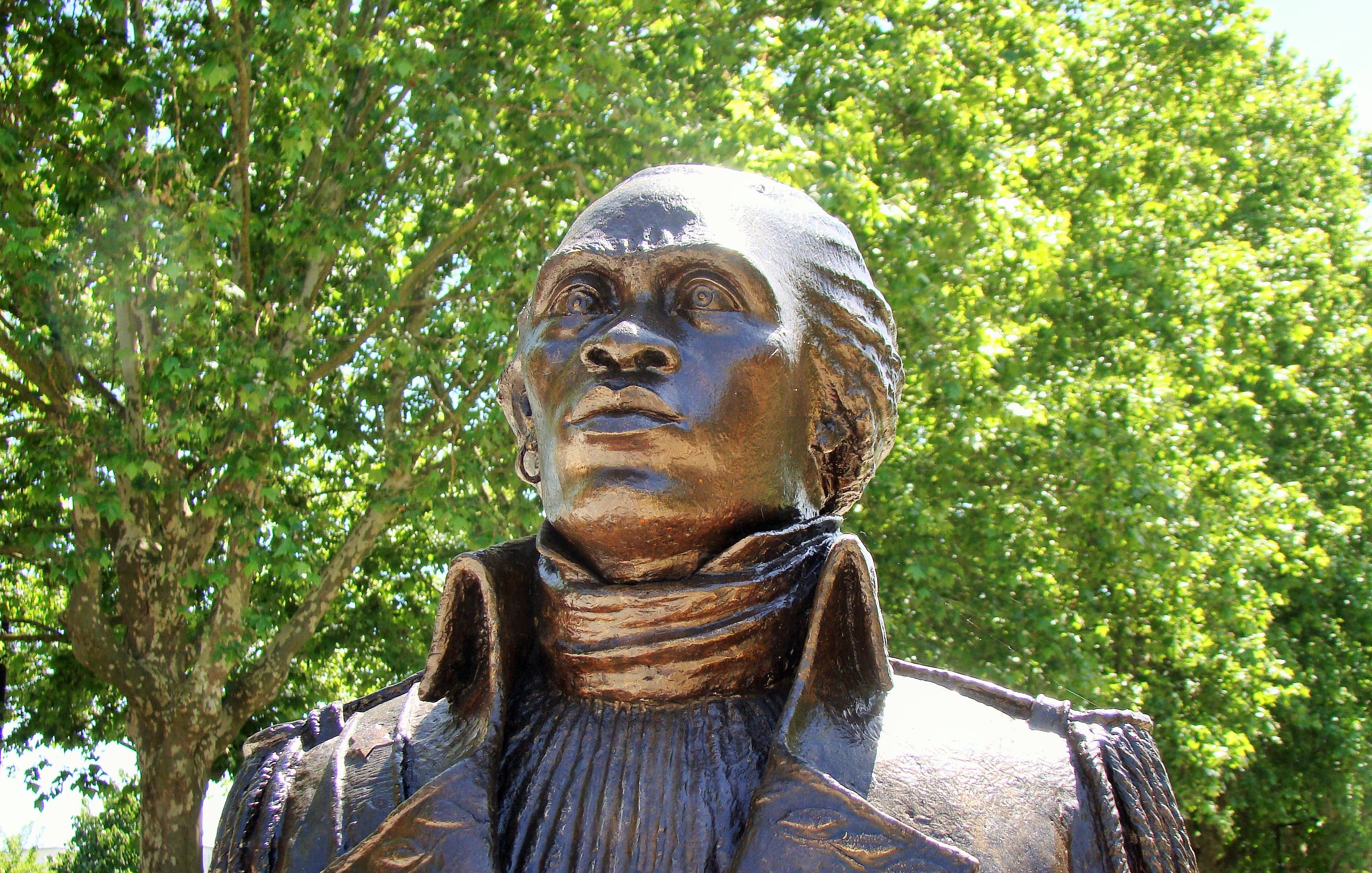 Voici la description de l'image : L'image montre une statue en bronze représentant un personnage en buste, probablement une figure historique. Le buste a un regard tourné vers le haut, suggérant une attitude déterminée ou réfléchie. Le personnage porte une veste à col haut, avec un plissé distinct sur le devant. Les détails du visage, bien que stylisés, montrent une expression forte, avec des traits bien définis. La statue est placée à l'extérieur, entourée d'arbres verdoyants, créant un cadre naturel agréable. La lumière du soleil illumine la statue, accentuant ses contours et sa texture.