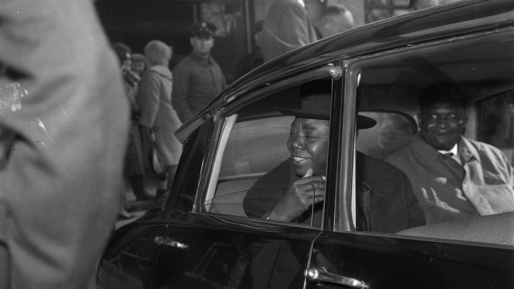 L'image montre l'intérieur d'une voiture noire, où deux hommes sont assis. L'un d'eux, qui se trouve à l'avant, semble souriant et engageant, habillé d'un costume élégant et portant un chapeau. Son expression indique qu'il est en train de parler ou de rire. À l'arrière de la voiture, l'autre homme a une expression plus neutre, vêtu également d'un manteau. À l'extérieur de la voiture, on peut apercevoir une foule de personnes en arrière-plan, certaines s'attardant dans un hall ou une gare d'arrivée. L'atmosphère semble animée, suggérant un moment de retrouvailles ou d'attente.