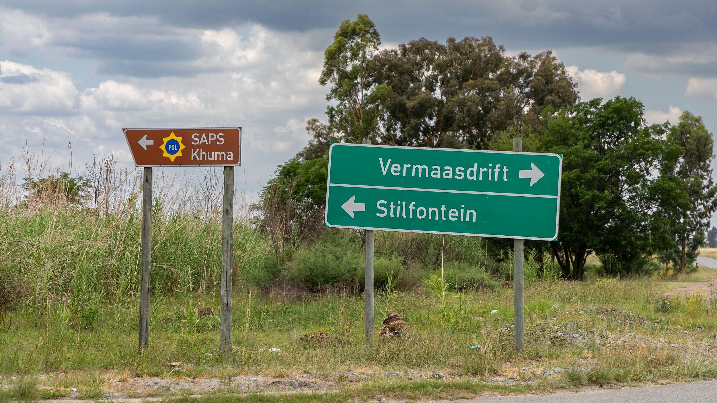 Cette image montre un carrefour routier en extérieur. À gauche, un panneau marron avec un symbole de police indique "SAPS Khuma", suggérant la présence d'un poste de police local. À droite, un panneau vert montre deux flèches directionnelles : une vers "Vermaasdrift" et l'autre vers "Stilfontein". L'environnement est caractérisé par une végétation verte et dense, avec quelques arbres en arrière-plan et un ciel nuageux, créant une atmosphère calme et naturelle. Le sol est asphalté, donnant l'impression d'une route peu fréquentée.