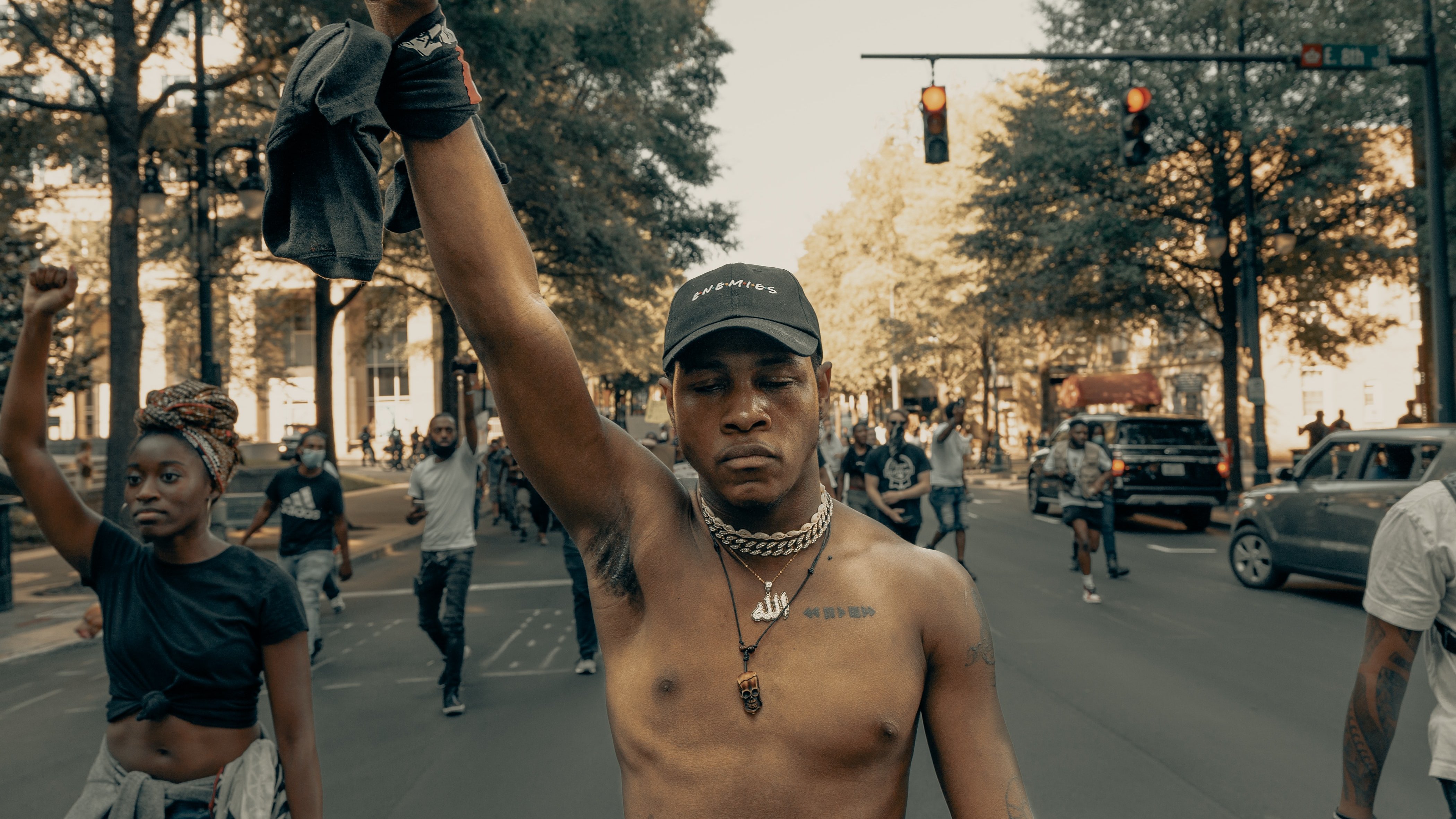 L'image montre une scène de manifestation dans une rue. Au premier plan, un jeune homme, torse nu, lève le poing en signe de protestation. Il porte une casquette et des chaînes autour du cou. À l'arrière-plan, d'autres personnes marchent, certaines levant également le poing, témoignant d'un esprit collectif. La rue est bordée d'arbres, et on peut apercevoir des voitures et des feux de signalisation. L'ambiance semble énergique et engagée, reflétant une lutte pour la justice ou l'égalité.
