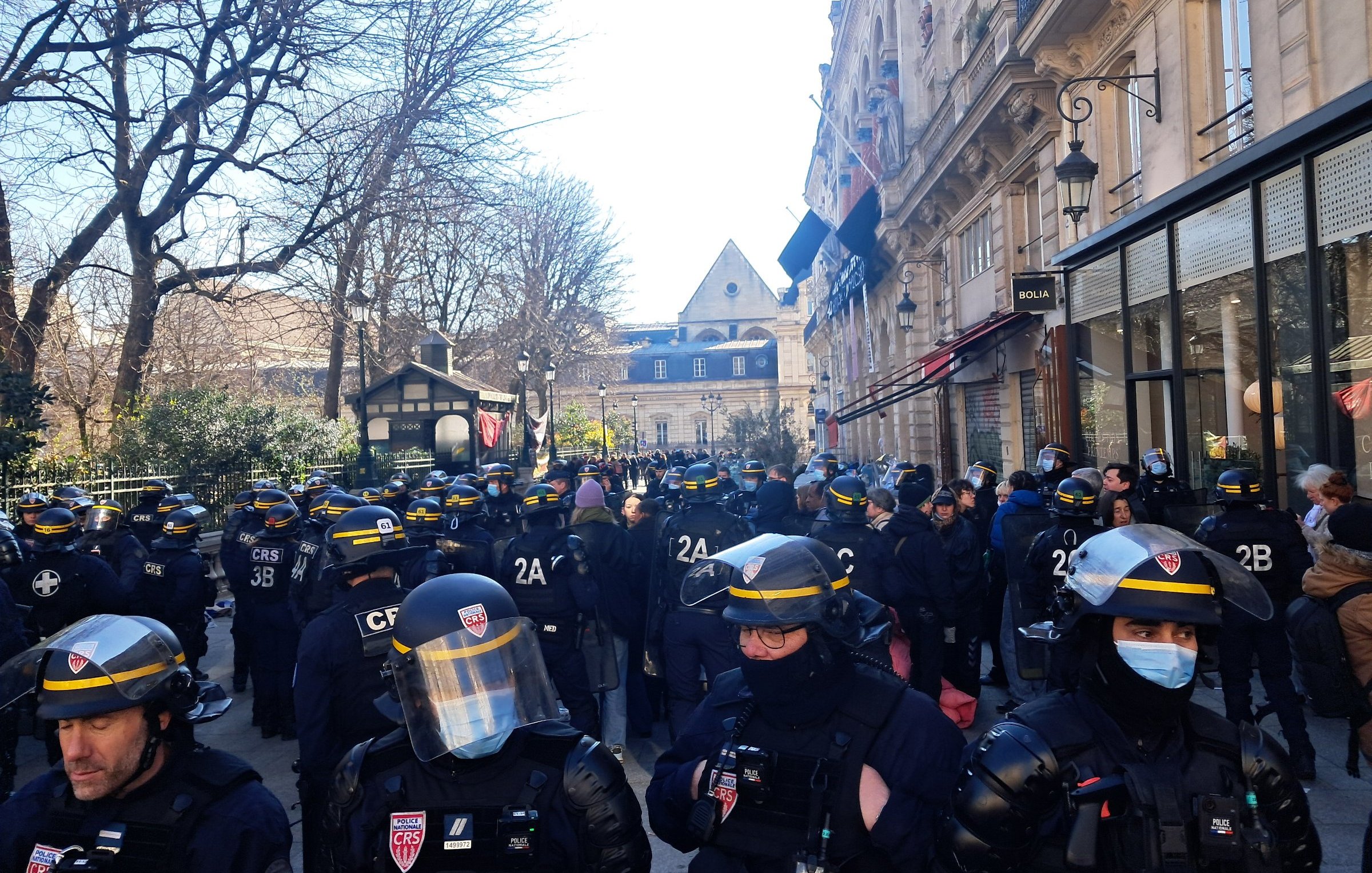 L'image montre une scène urbaine animée. En premier plan, un groupe de policiers en uniforme, équipés de casques et de protections, se tient rassemblé. Leur posture est attentive, suggérant une opération en cours ou une manifestation. À l'arrière-plan, des arbres dénudés et des bâtiments historiques sont visibles, indiquant que la scène se déroule probablement dans une zone urbaine. C'est une journée ensoleillée, avec un ciel clair, apportant une lumière vive sur la scène. L'atmosphère semble tendue, avec des gens qui observent la situation depuis les côtés. On peut percevoir une impression d'encadrement, de sécurité, mais aussi de tension sociale.