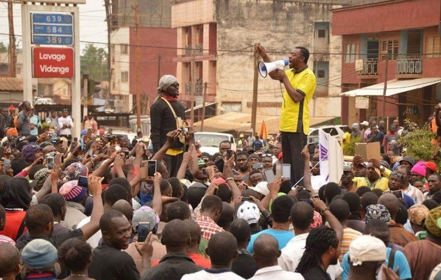 L'image montre une scène de rassemblement dans une rue animée, remplie de nombreuses personnes. Au centre, un orateur, habillé en jaune, se tient sur une plateforme surélevée, tenant un mégaphone et s'adressant à la foule. Les membres de l'audience, qui semblent nombreux et engagés, lèvent souvent leurs téléphones, capturant probablement des moments du discours. En arrière-plan, on aperçoit des bâtiments et une affichage indiquant des prix, ce qui suggère un environnement urbain. L'atmosphère est énergique, marquée par l'enthousiasme et la mobilisation des participants.