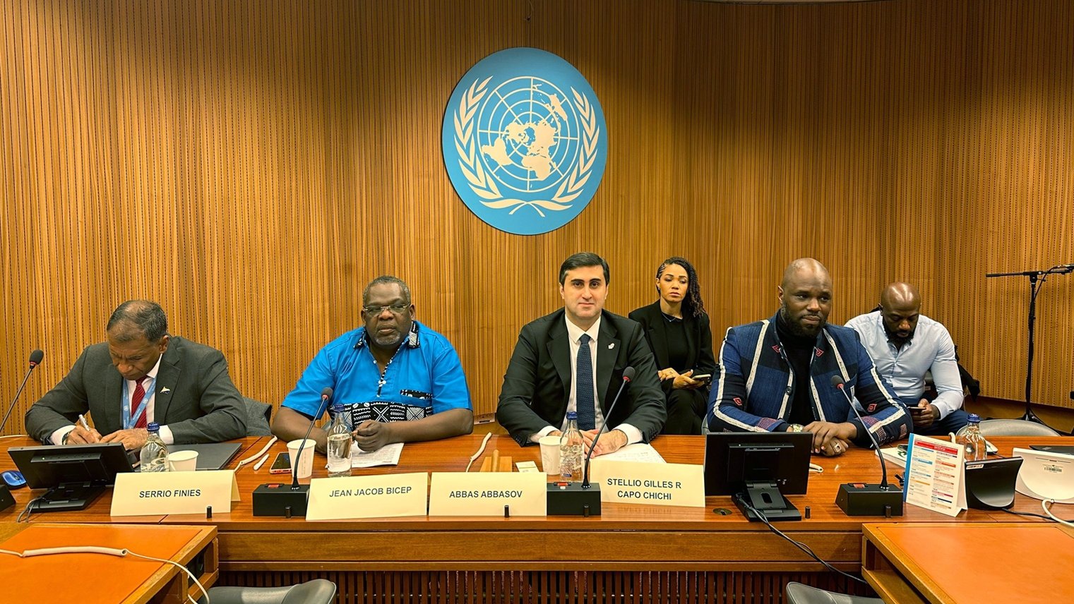 L'image montre une scène à une table de conférence, probablement lors d'une réunion aux Nations Unies. Au centre, un homme en costume noir et cravate blanche, avec un air sérieux, semble être le porte-parole ou le modérateur. À sa gauche, un homme portant une chemise bleue et des lunettes, avec un air attentif. À sa droite, un homme d'apparence plus décontractée avec un pull et une barbe, qui semble également participer à la discussion. En arrière-plan, plusieurs personnes sont assises, écoutant attentivement. Le décor est sobre, avec des murs en bois et le logo des Nations Unies en arrière-plan.