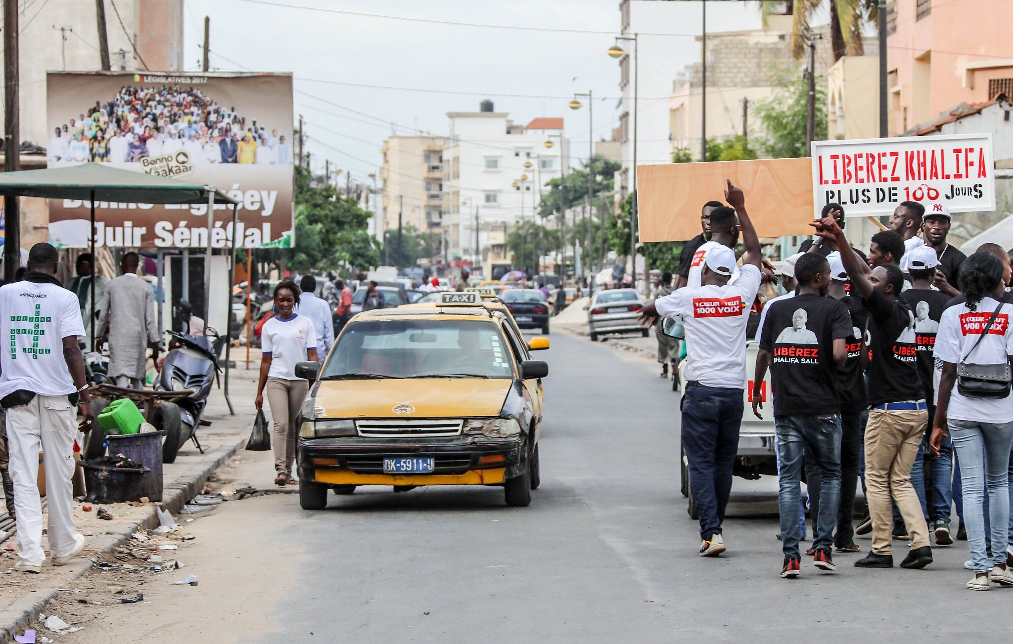 L'image montre une scène animée dans une rue urbaine. À gauche, des personnes marchent, certaines portant des tee-shirts blancs avec des inscriptions visibles. Ils semblent participer à une manifestation ou un rassemblement. Au centre, un taxi jaune est garé sur le côté de la route. Sur le côté droit, il y a plusieurs personnes, et l'ambiance donne une impression de dynamisme et d'engagement. Des affiches sont visibles en arrière-plan, dont l'une mentionne des slogans politiques, suggérant un contexte de revendication ou de mobilisation sociale. Le bruit de la ville, les voix des manifestants et le bruit des véhicules complètent cette atmosphère vivante.