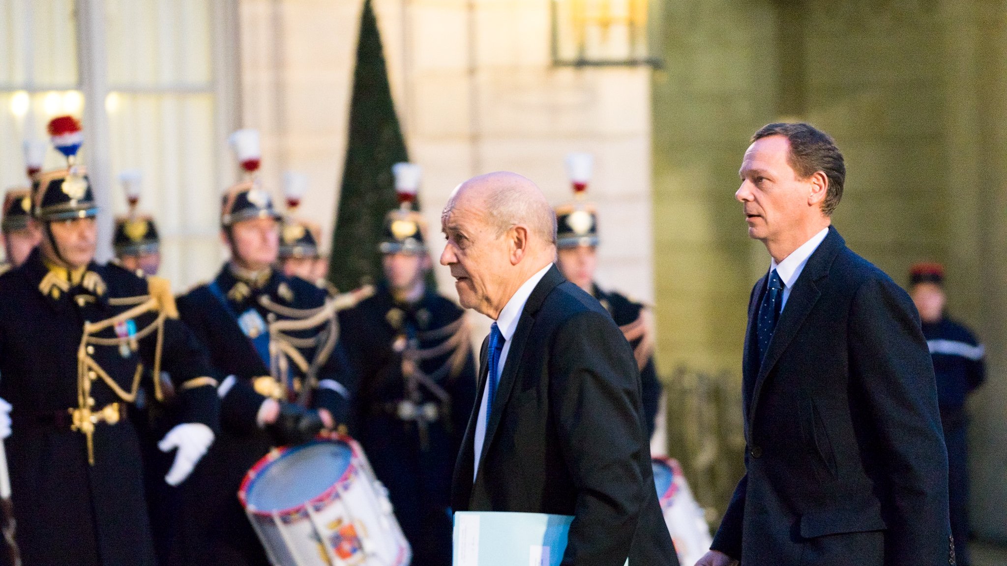L'image montre deux hommes marchant dans une ambiance officielle. À l'arrière-plan, on peut apercevoir une garde cérémonielle, avec des soldats en uniforme traditionnel, portant des chapeaux distinctifs et tenant des instruments de musique, comme des tambours. Les hommes au premier plan semblent se rendre à une réunion importante, l'un d'eux portant un dossier ou un document. L'éclairage de la scène est doux, créant une atmosphère solennelle et respectueuse.