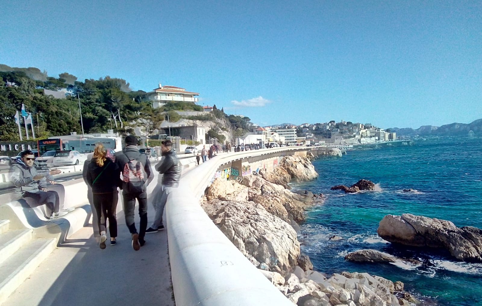 Imaginez-vous sur une promenade longeant la mer. Sur votre droite, vous pouvez entendre le bruit des vagues qui s'écrasent doucement contre les rochers, tandis que l'odeur salée de l'océan flotte dans l'air. Le vent léger caresse votre visage et apporte une sensation de fraîcheur. À votre gauche, des bâtiments se dressent, certains sont des maisons et d'autres des commerces. Vous pouvez sentir l'animation de la ville avec des groupes de personnes qui marchent, certains discutant joyeusement. La promenade est large et bien aménagée, invitant à la flânerie. Au loin, le paysage est ponctué d'arbres et de collines, avec quelques nuages blancs dégradant le bleu du ciel. La lumière du soleil illumine tout le paysage, créant une ambiance chaleureuse. L'ensemble évoque une belle journée au bord de la mer, propice à la détente et au plaisir des sens.