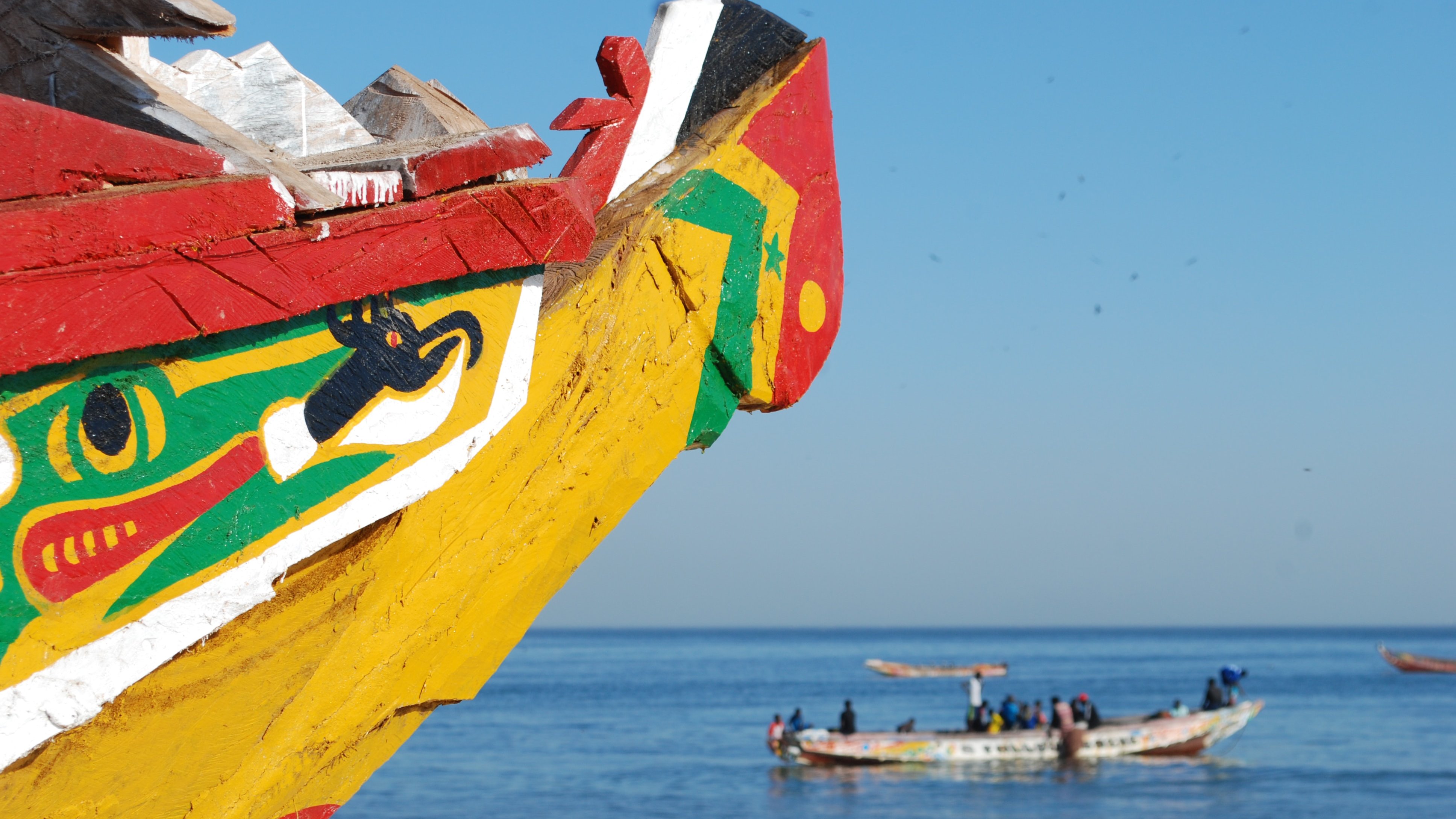 Cette image représente la proue colorée d'une pirogue traditionnelle, peinte de motifs vifs dans des couleurs comme le rouge, le jaune, et le vert. Au loin, on peut apercevoir d'autres bateaux de pêche qui naviguent paisiblement sur l'eau calme d'une mer bleue. Le ciel est dégagé, créant une atmosphère sereine et ensoleillée. L'air salin doit être frais, et on peut entendre le bruit des vagues et peut-être des conversations des marins qui travaillent sur leurs embarcations. L'ensemble dégage une ambiance maritime typique des côtes africaines.
