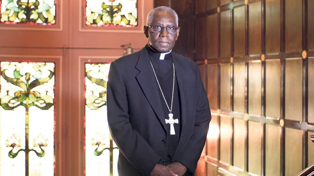 Dans cette image, nous voyons un homme debout dans un environnement qui semble être une pièce élégante, probablement une église ou un bureau religieux. Il porte une tenue noire, typique des membres du clergé, avec une soutane et un col ecclésiastique. Autour de son cou, une grande croix pend, symbolisant son rôle religieux. Le fond est composé de murs en bois foncé et de vitraux colorés, ce qui crée une atmosphère solennelle et respectueuse. Les motifs des vitraux laissent transparaître une lumière douce, ajoutant une dimension spirituelle à l'ensemble de la scène. L'homme a une expression sérieuse, se tenant droit et digne, ce qui évoque l'autorité et la sagesse.