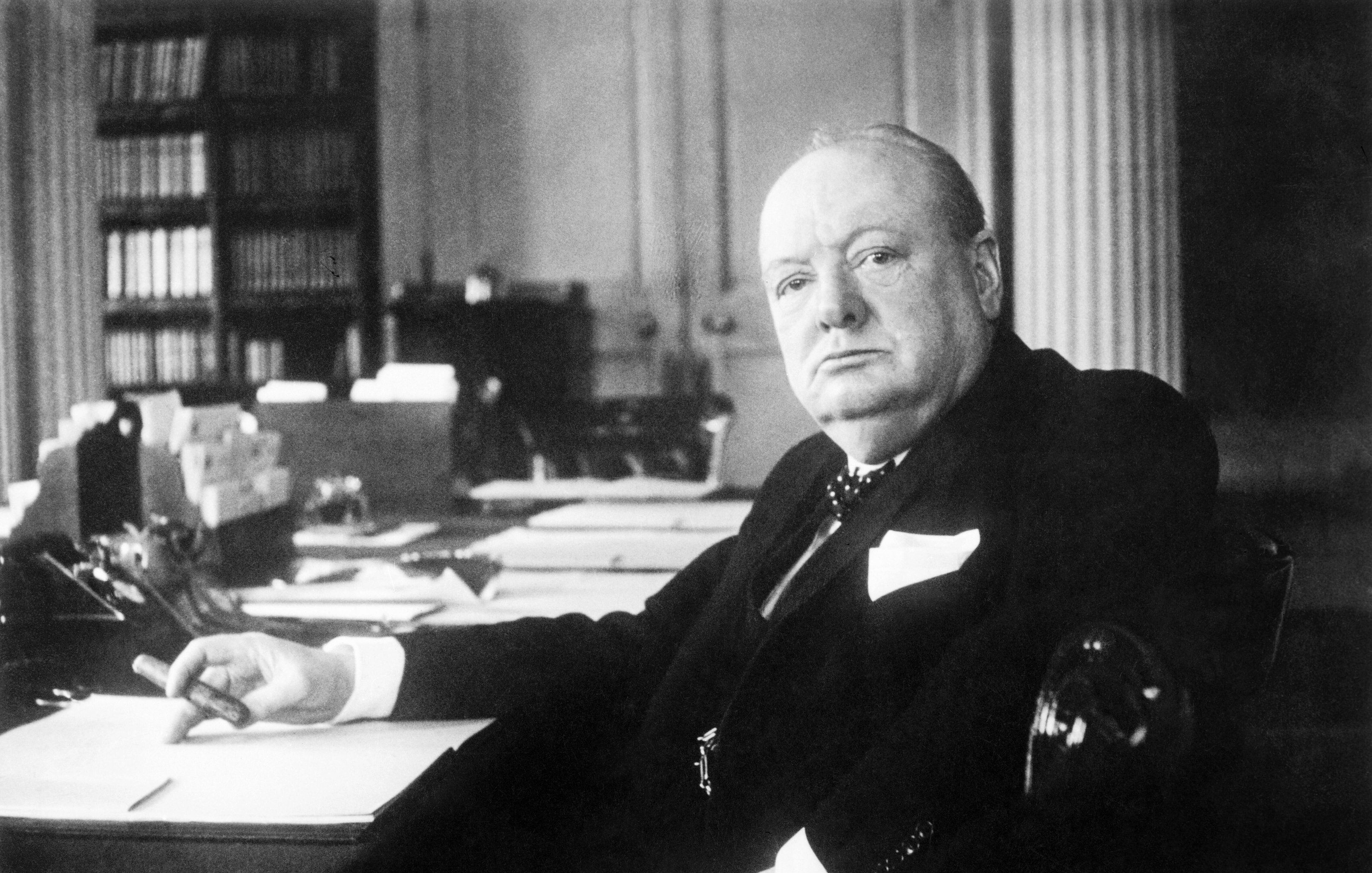 L'image présente un homme assis à un bureau. Il a une expression sérieuse et regarde droit devant lui. Son visage est rond et bien en chair, avec des traits marqués. Il est vêtu d'un costume sombre, avec une chemise claire et une cravate. Dans sa main, il tient un stylo, et devant lui, des feuilles de papier sont disposées sur le bureau. En arrière-plan, on distingue des étagères remplies de livres. L'atmosphère de l'image suggère une ambiance de travail studieuse et normalement formelle.