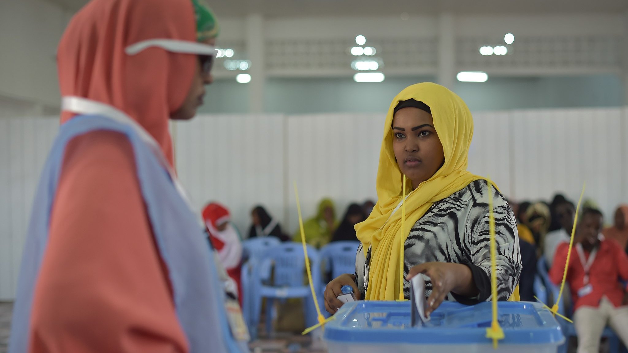 L'image montre une scène de vote dans un lieu organisé. À gauche, une femme porte un hijab orange et des lunettes de soleil, regardant vers une autre femme, qui est située à droite. Cette dernière, en hijab jaune, semble concentrée alors qu'elle dépose son bulletin dans une urne transparente devant elle. L'intérieur de la salle est éclairé, avec des chaises en plastique bleues disposées en rangées. On peut percevoir une atmosphère de participation et d'engagement civique. Des femmes en arrière-plan sont également présentes, créant une ambiance communautaire.