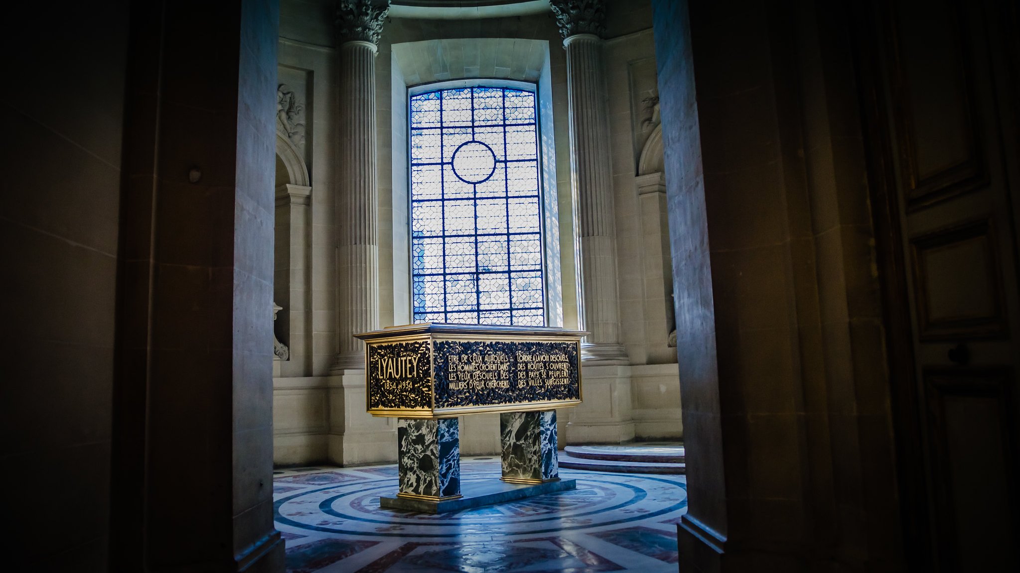 Cette image montre une pièce d'un bâtiment majestueux, probablement un musée ou un palais. Au centre, se trouve un coffre sombre et décoré, surélevé sur un socle circulaire. Les côtés du coffre sont ornés de motifs complexes et d'inscriptions dorées. En arrière-plan, une grande fenêtre ornée de vitraux diffuse une lumière douce qui éclaire la pièce. Les murs sont en pierre grise, enrichis de colonnes majestueuses. L'atmosphère générale est à la fois calme et solennelle, invitant à la contemplation.