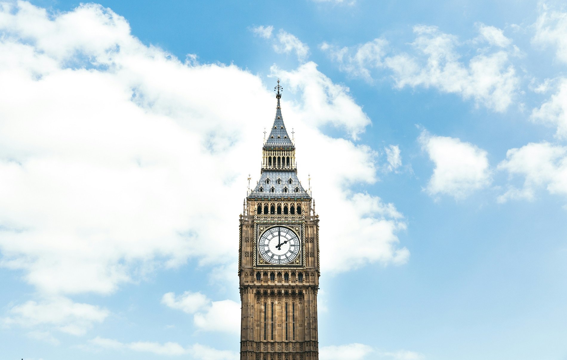 Imaginez un grand clocher en pierre, qui s'élève majestueusement vers le ciel. Ce bâtiment emblématique, connu sous le nom de Big Ben, est orné d'une grande horloge dont les aiguilles sont parfaitement visibles, indiquant l'heure avec précision. La tour est décorée de détails architecturaux, avec des voûtes et des motifs, et se dresse dans un ciel bleu parsemé de nuages blancs cotonneux. La lumière du jour accentue les couleurs chaudes de la pierre, ajoutant une touche rayonnante à cette structure historique.