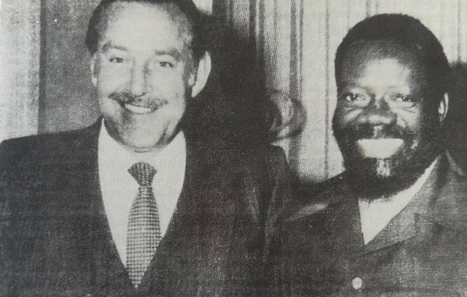 L'image montre deux hommes se tenant côte à côte, souriant largement. Le premier homme, à gauche, porte un costume sombre avec une cravate. Il a des cheveux grisonnants, une moustache bien dessinée et une expression amicale. Le deuxième homme, à droite, est habillé d'une veste de style militaire et a une barbe bien fournie. Il a les cheveux frisés et sourit également, ce qui donne une atmosphère chaleureuse à la photo. Les deux tiennent des verres à la main, suggérant une occasion conviviale, peut-être une célébration ou un événement. L'arrière-plan est flou, mettant l'accent sur les deux hommes.