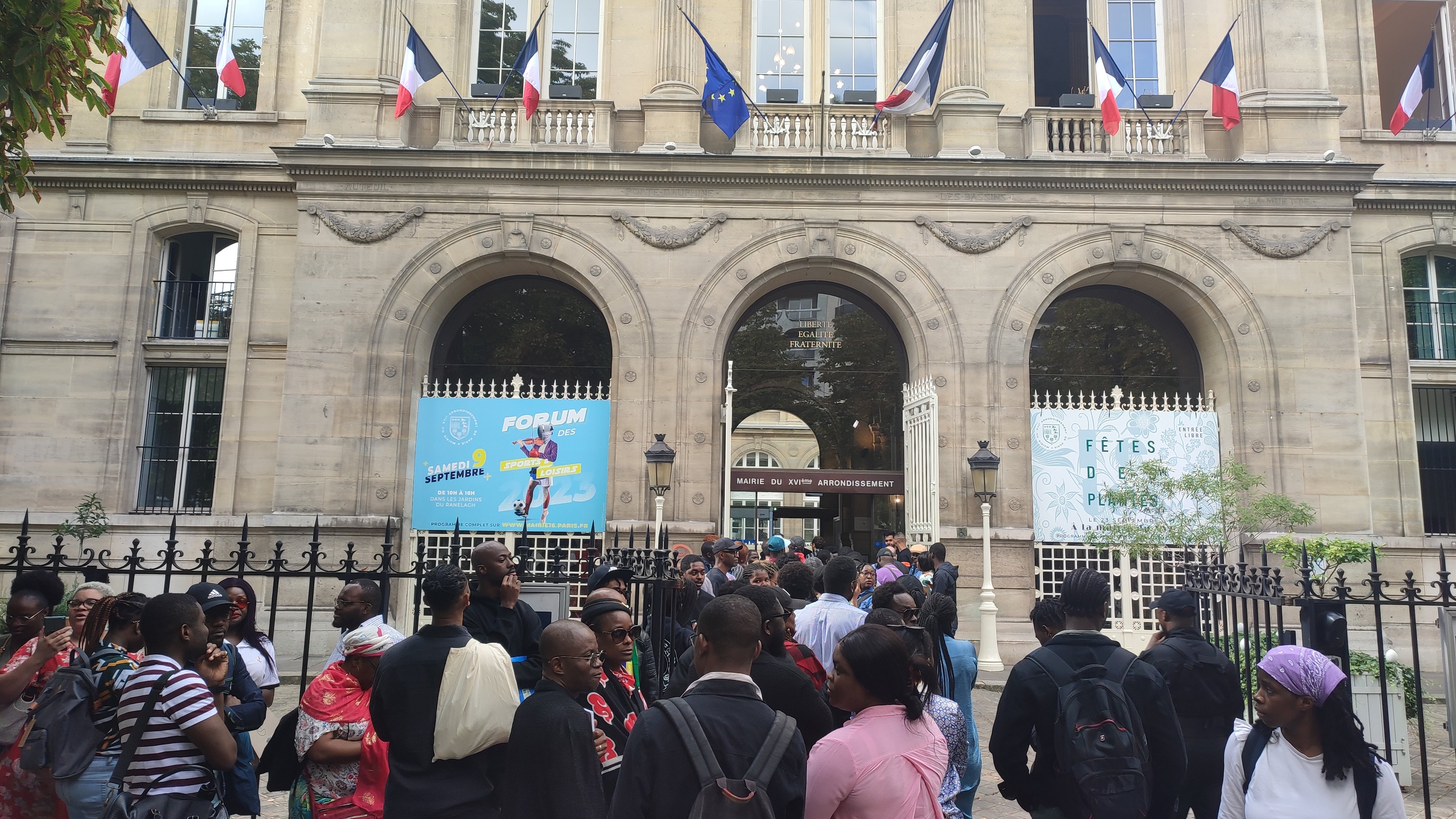 L'image montre un bâtiment imposant avec une architecture classique, visible grâce à ses grandes fenêtres et ses arches. Devant ce bâtiment, un groupe de personnes se rassemble, créant une atmosphère animée. Des drapeaux français flottent au-dessus d'eux, témoignant de l'importance de ce lieu. Les visiteurs semblent variés en termes d'âge et de style vestimentaire, ce qui reflète une diversité culturelle. À l'arrière-plan, des affiches colorées annoncent un événement, ajoutant une touche festive à la scène. L'ensemble dégage une ambiance d'attente et d'excitation.