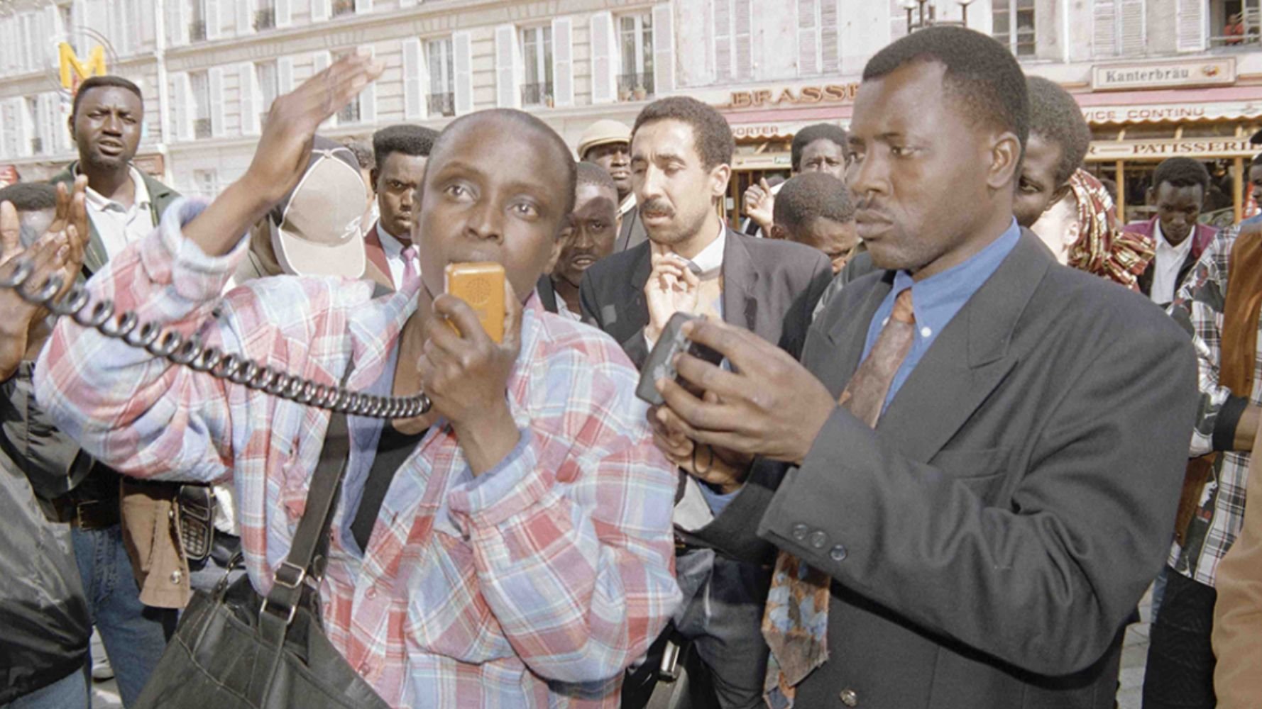 L'image montre un groupe de personnes rassemblées dans un espace public animé. Au premier plan, deux hommes attirent l'attention. L'un d'eux, portant une chemise à carreaux et tenant un microphone, semble s'exprimer avec passion, les bras levés, signalant une forte émotion. L'autre homme, en costume sombre, tient un appareil photo, observant et capturant le moment. En arrière-plan, on peut apercevoir une foule diverse, des visages attentifs, écoutant ou réagissant à la situation. L'atmosphère générale est dynamique, avec un sentiment de mobilisation ou de protestation.