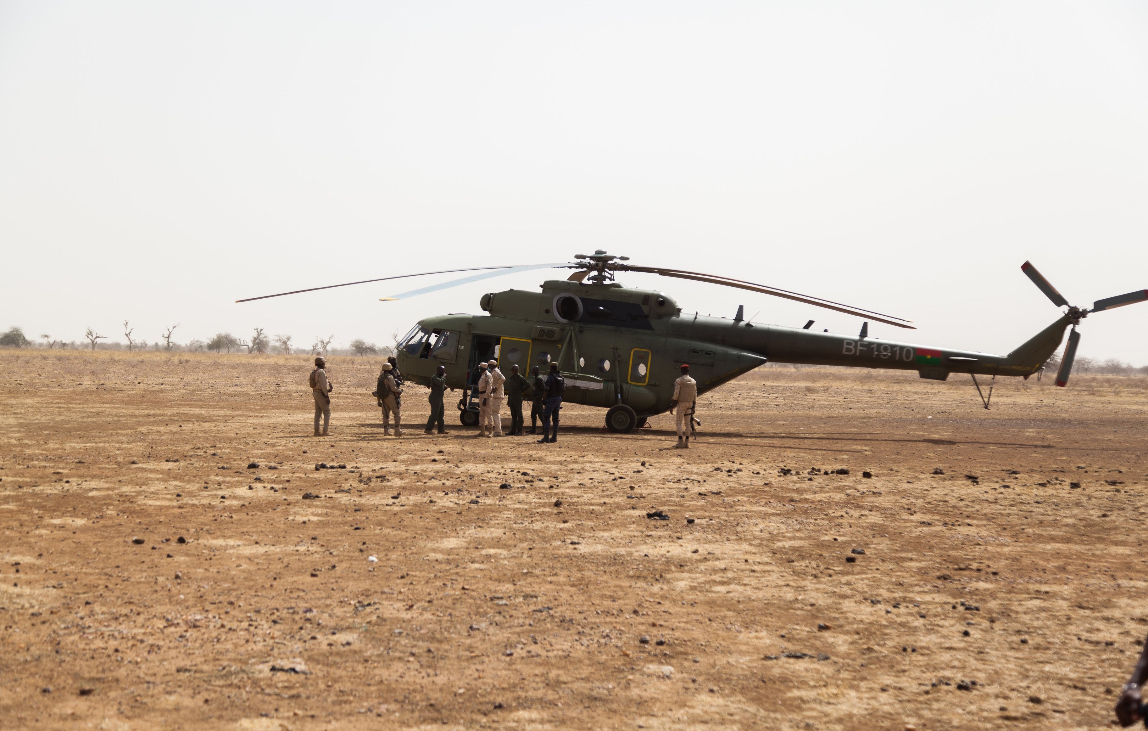 L'image montre un hélicoptère posé sur un terrain désertique, avec un sol caillouteux et sec. Autour de l'hélicoptère, plusieurs personnes sont rassemblées, certaines en uniforme militaire. La scène semble se dérouler dans une région aride, avec peu de végétation en arrière-plan.